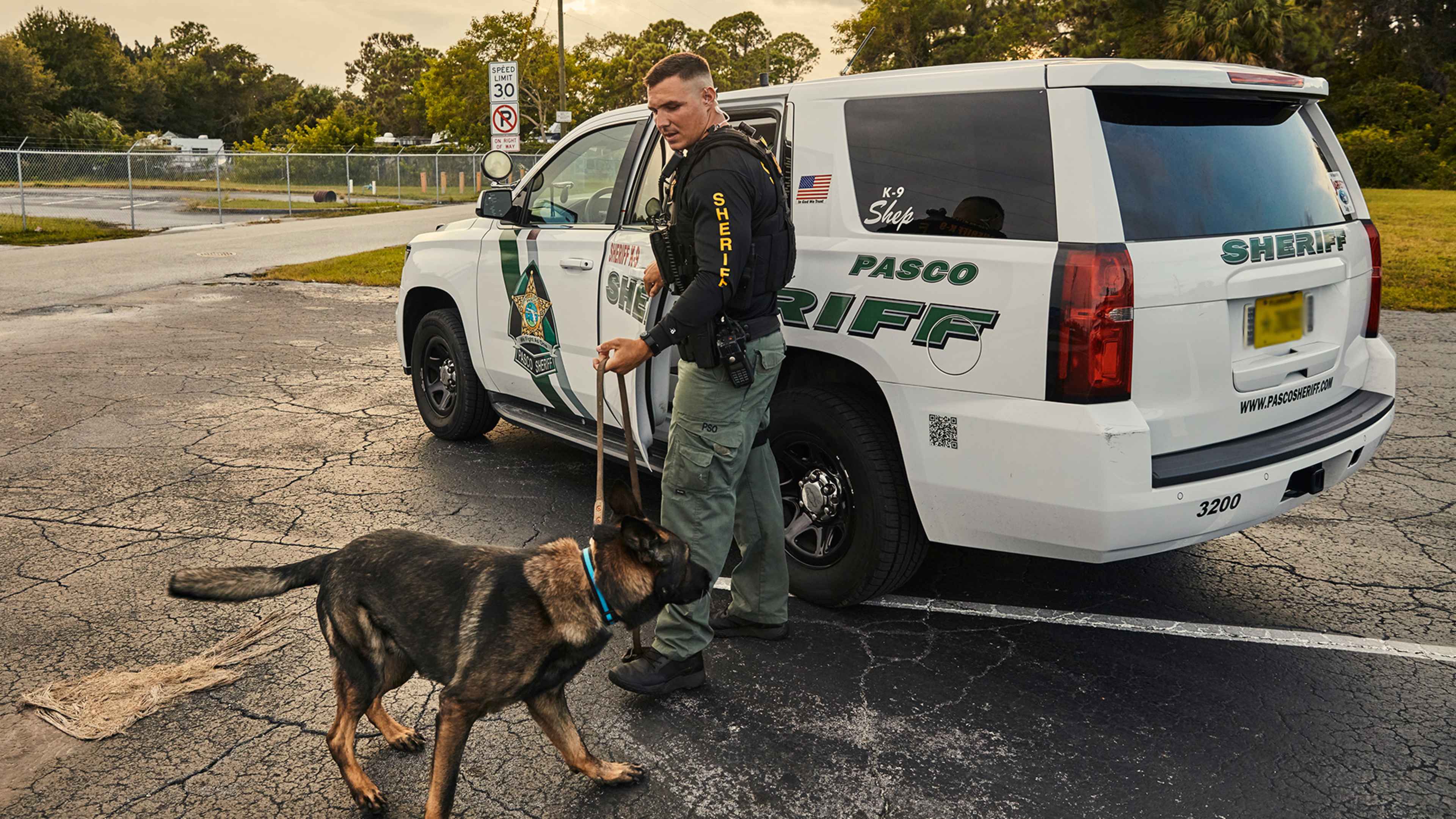 A law enforcement officer stands next to a police vehicle, with a K-9 dog at their side, in an outdoor setting with trees and a road visible in the background.
