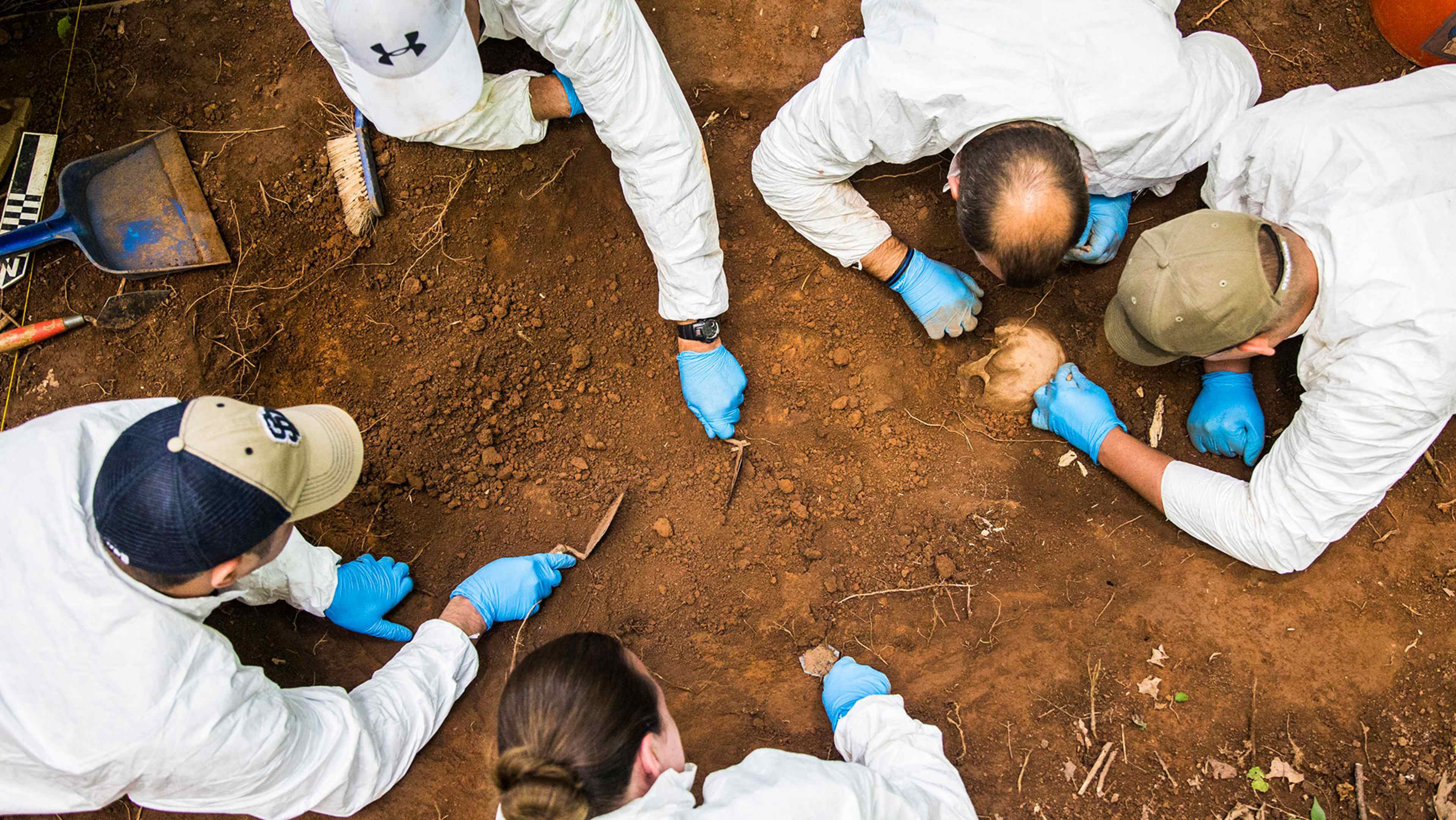 The image shows several people in protective white suits examining a dirt-covered area, likely conducting an investigation or analysis of some kind.