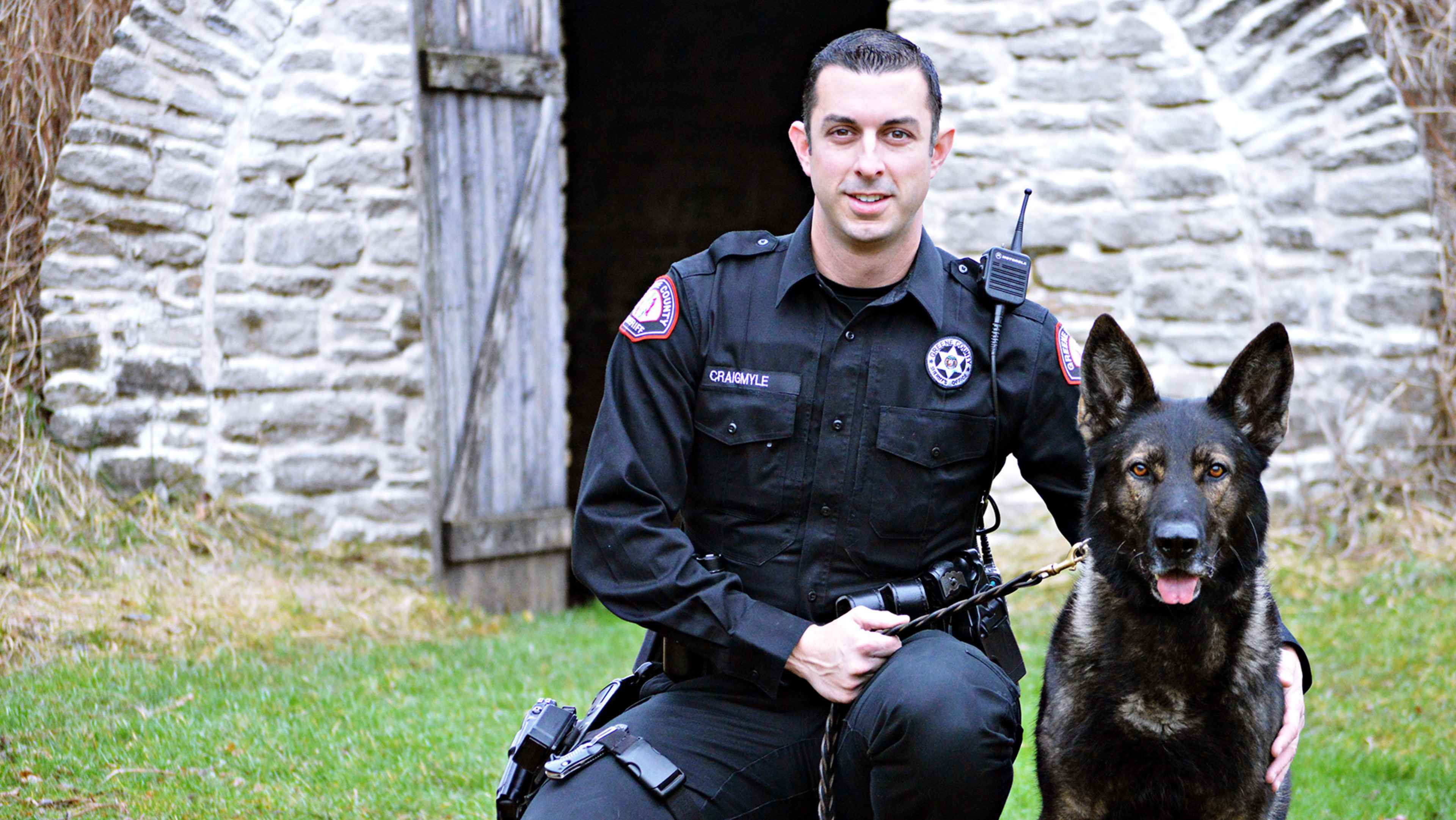 A uniformed police officer stands in a grassy field, accompanied by a German shepherd dog, in front of a stone building.