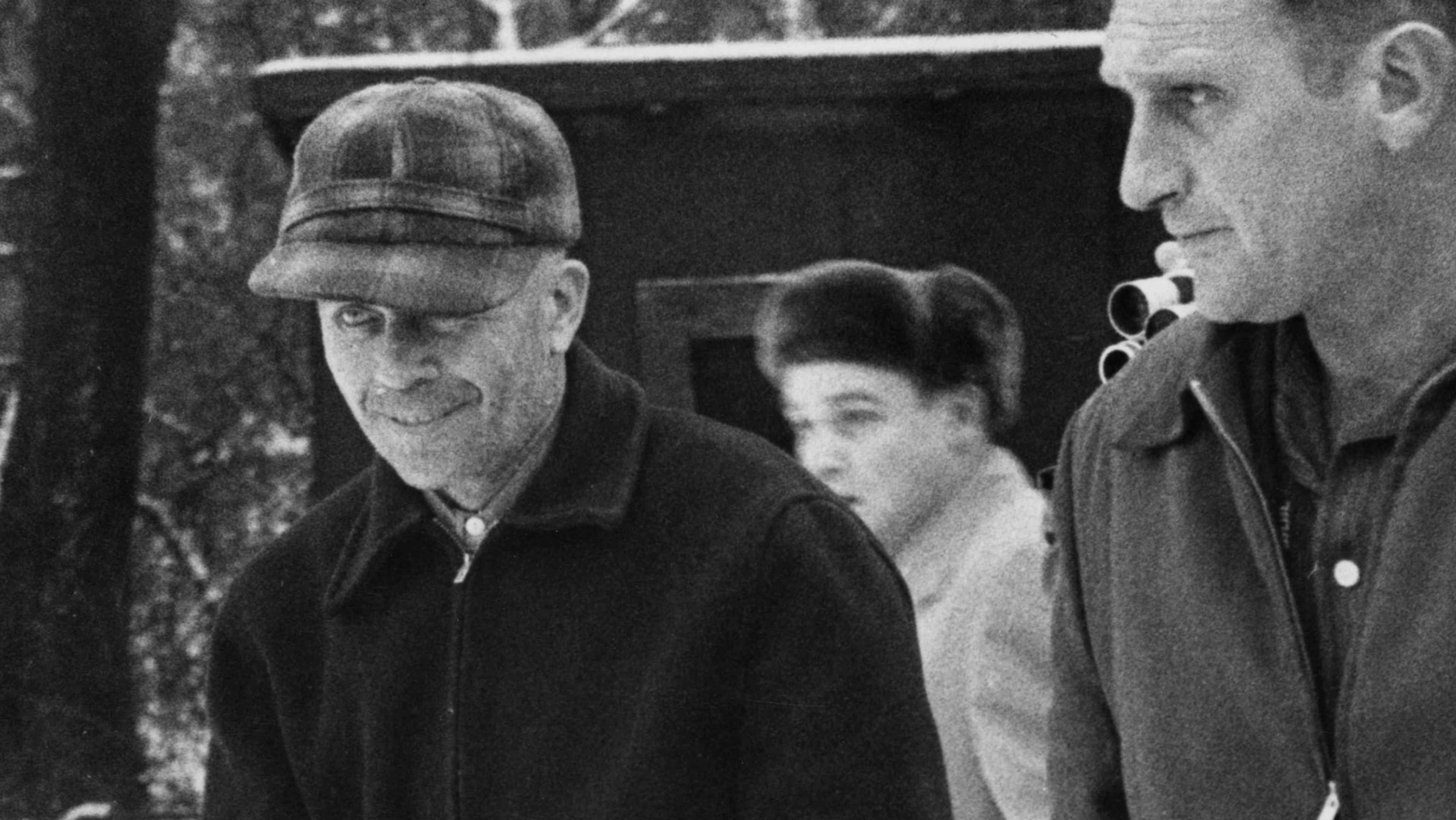 The image depicts three individuals in a black and white photograph. The foreground shows an older man wearing a cap, while behind him are two other people, one of whom appears to be a woman.
