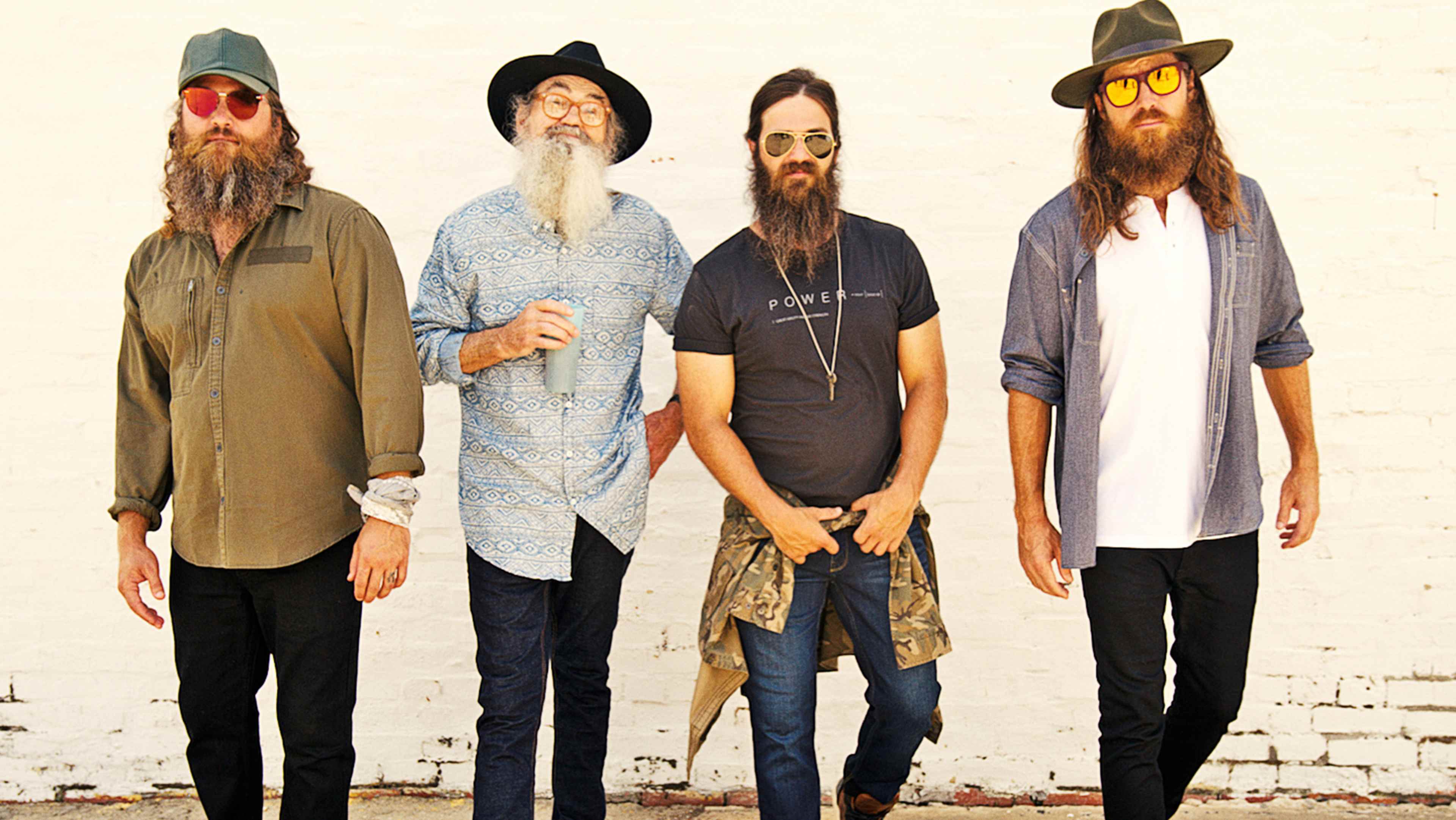 Four men with long beards and distinctive fashion styles, standing in front of a plain white wall.