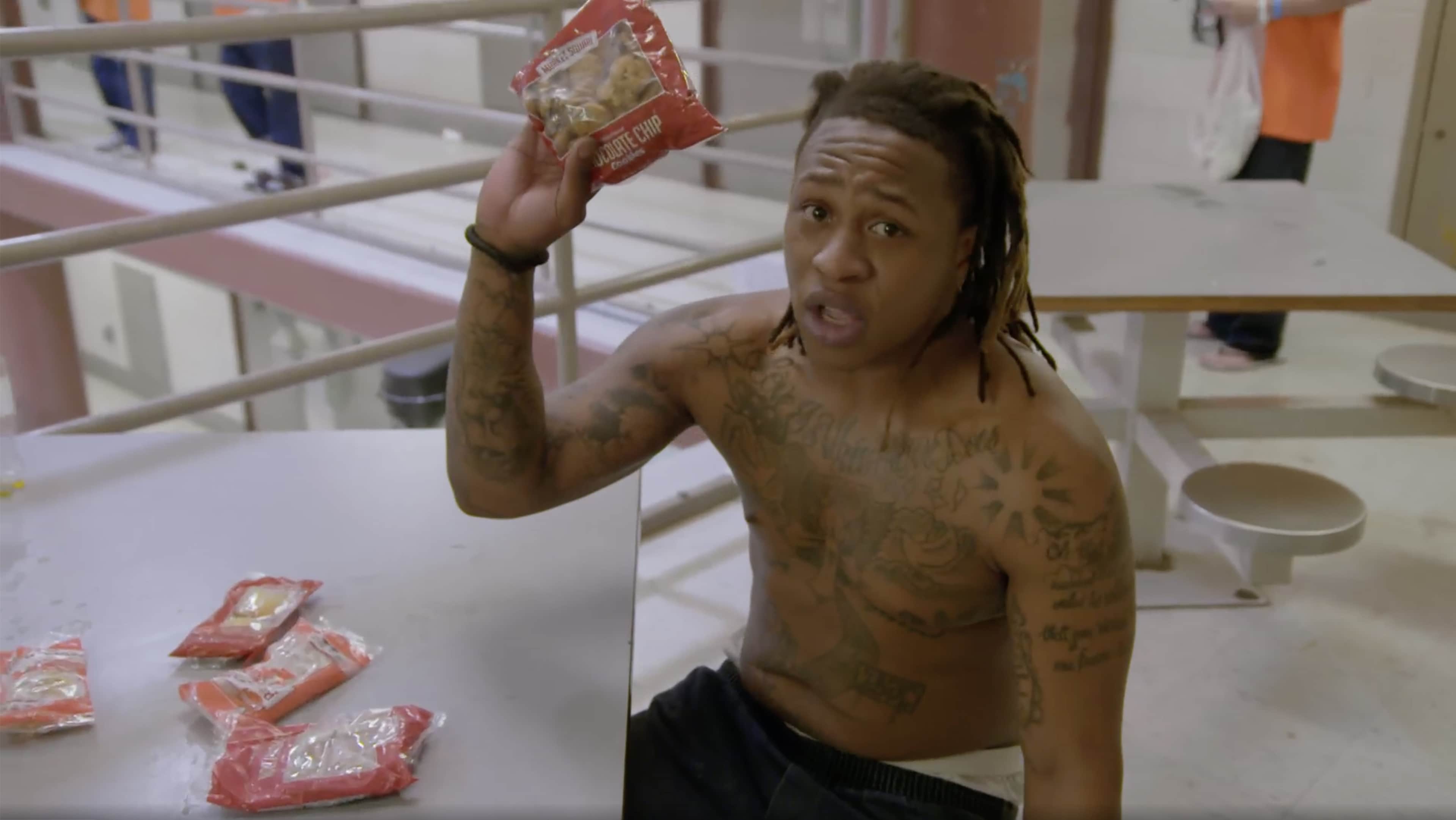 A shirtless individual with tattoos on their body is holding a snack in their hand, standing in what appears to be a hospital or medical facility setting.