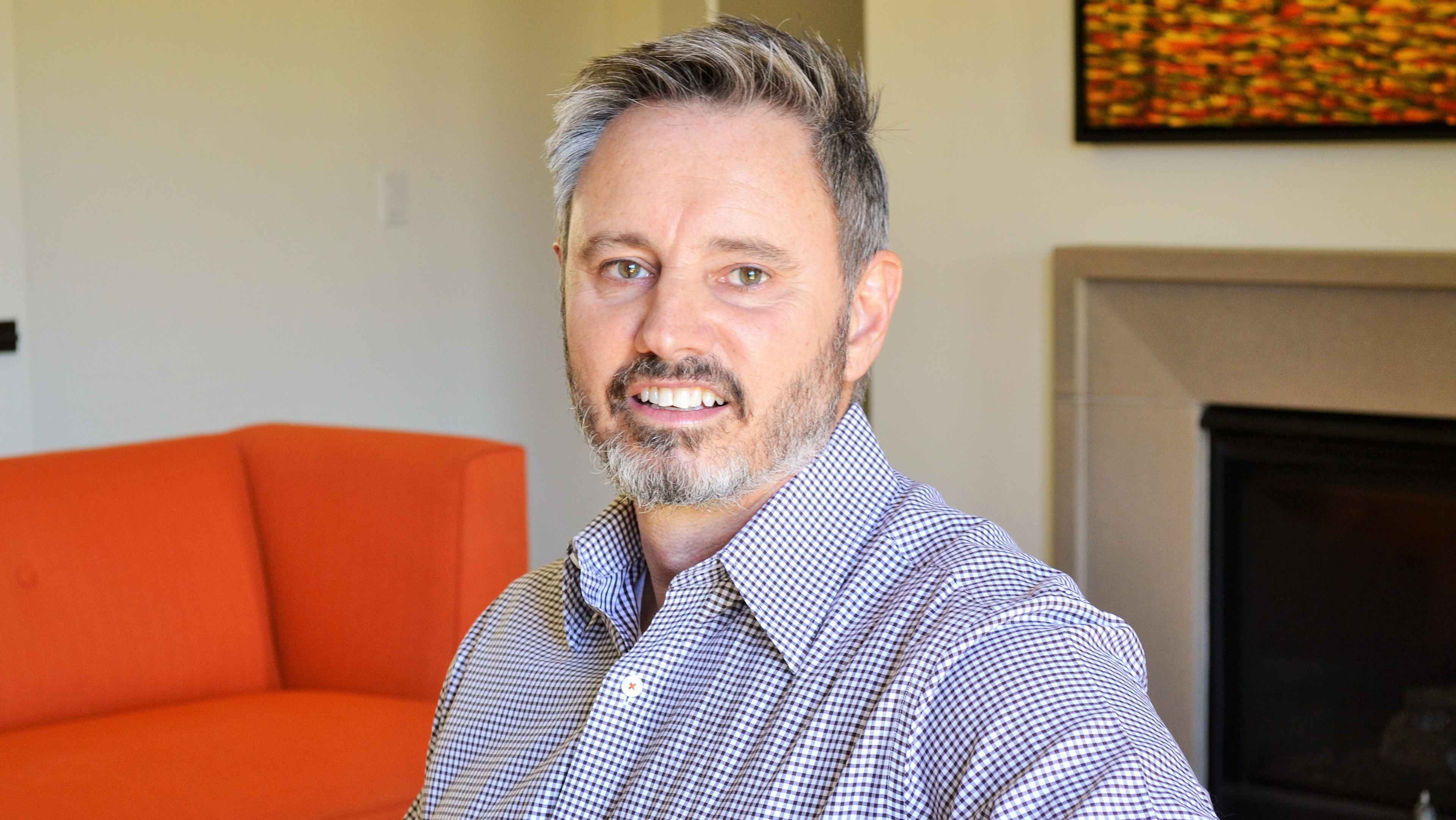 The image shows a middle-aged man with gray hair and a beard, wearing a checkered shirt, sitting on an orange couch in what appears to be a living room setting with a framed artwork on the wall behind him.