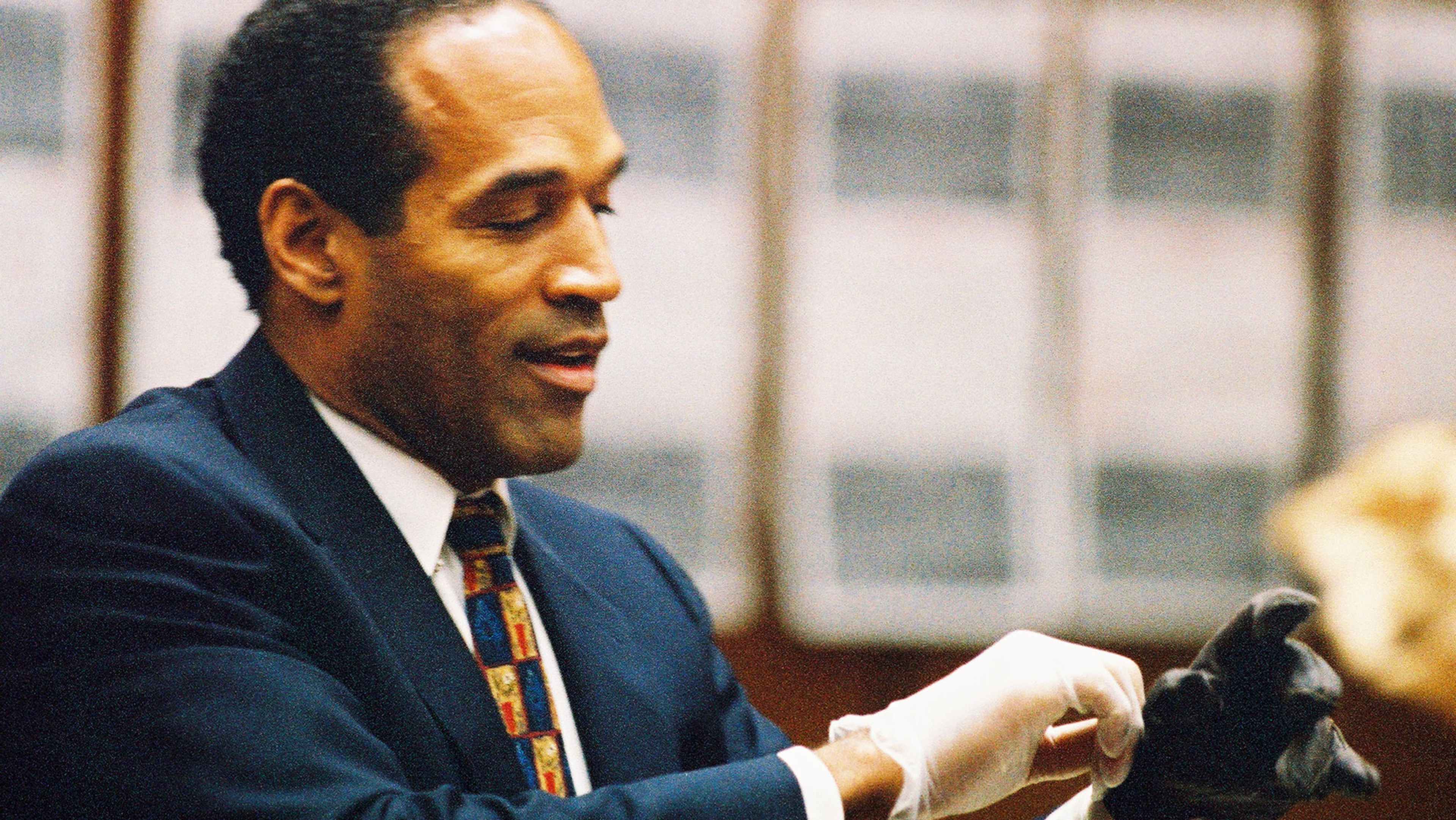A man in a dark suit and tie is sitting at a desk, with a window visible in the background.