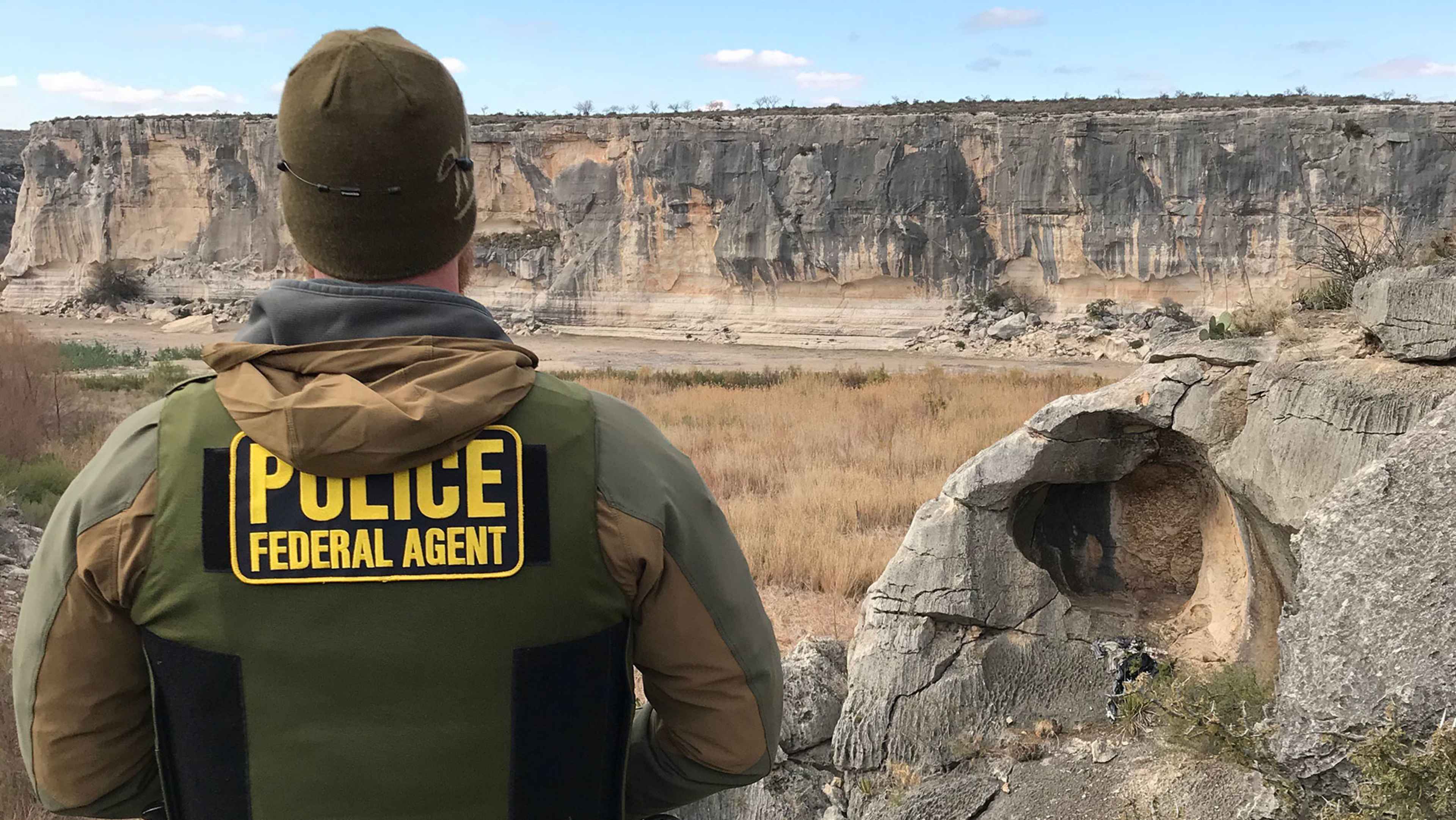 A federal agent in a police vest stands in the foreground, facing a vast, rugged landscape of towering cliffs and rock formations in the background.