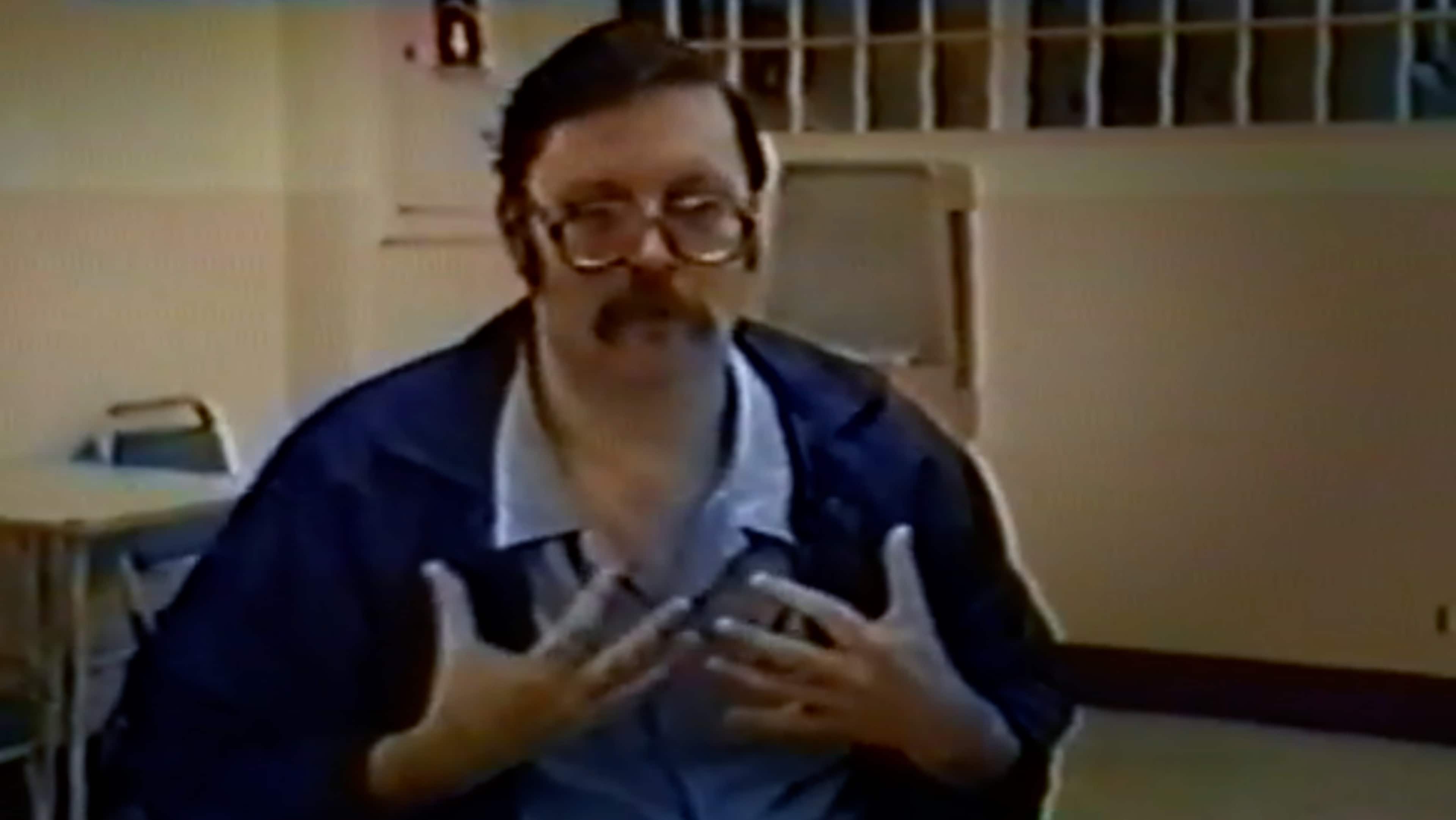 The image shows a man with a beard and glasses, wearing a blue jacket, sitting at a table and holding some papers in his hands.