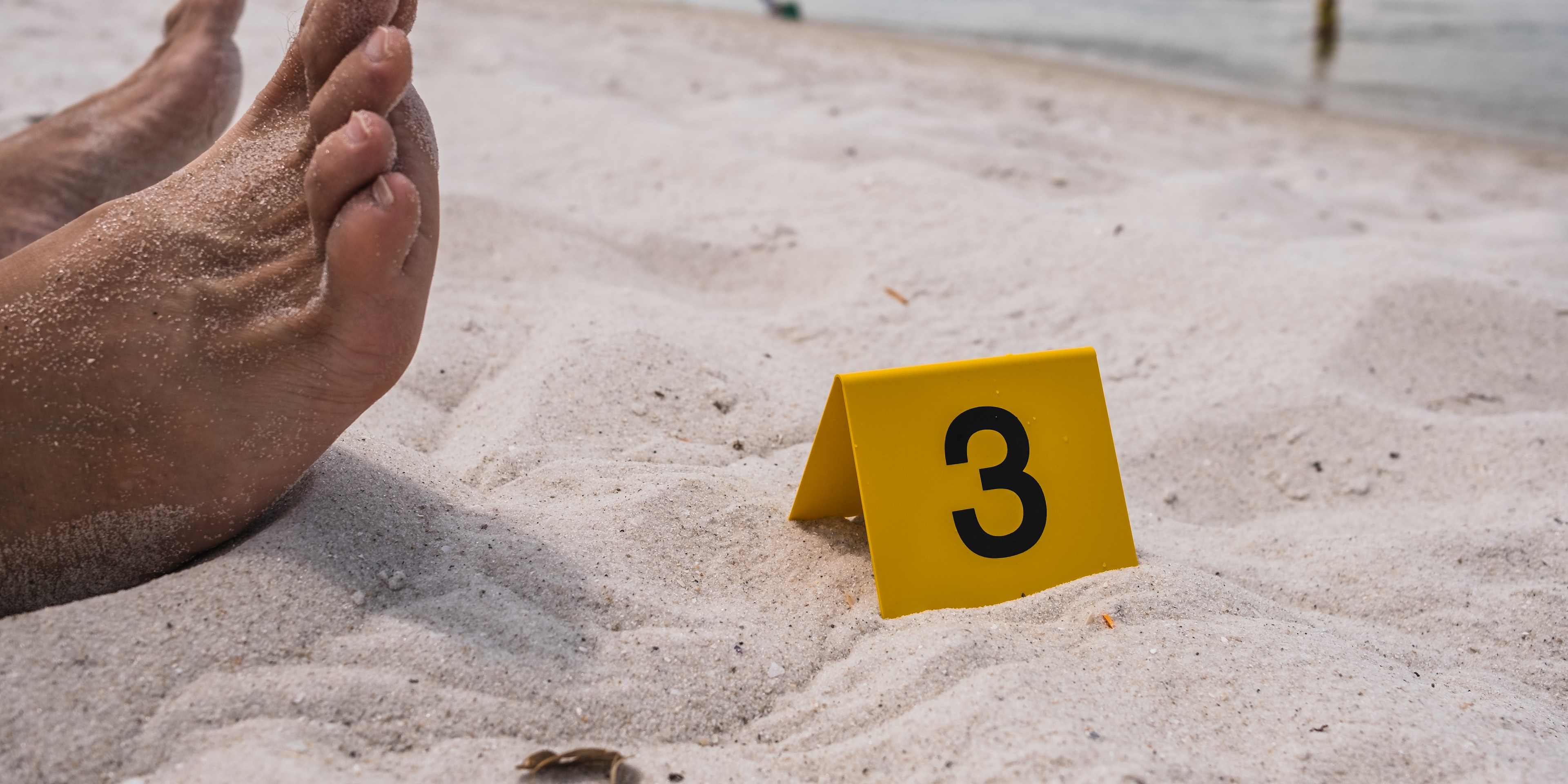 One yellow crime scene evidence marker next to the foot of a corpse laying on the beach after a murder shooting stabbing body forensics csi