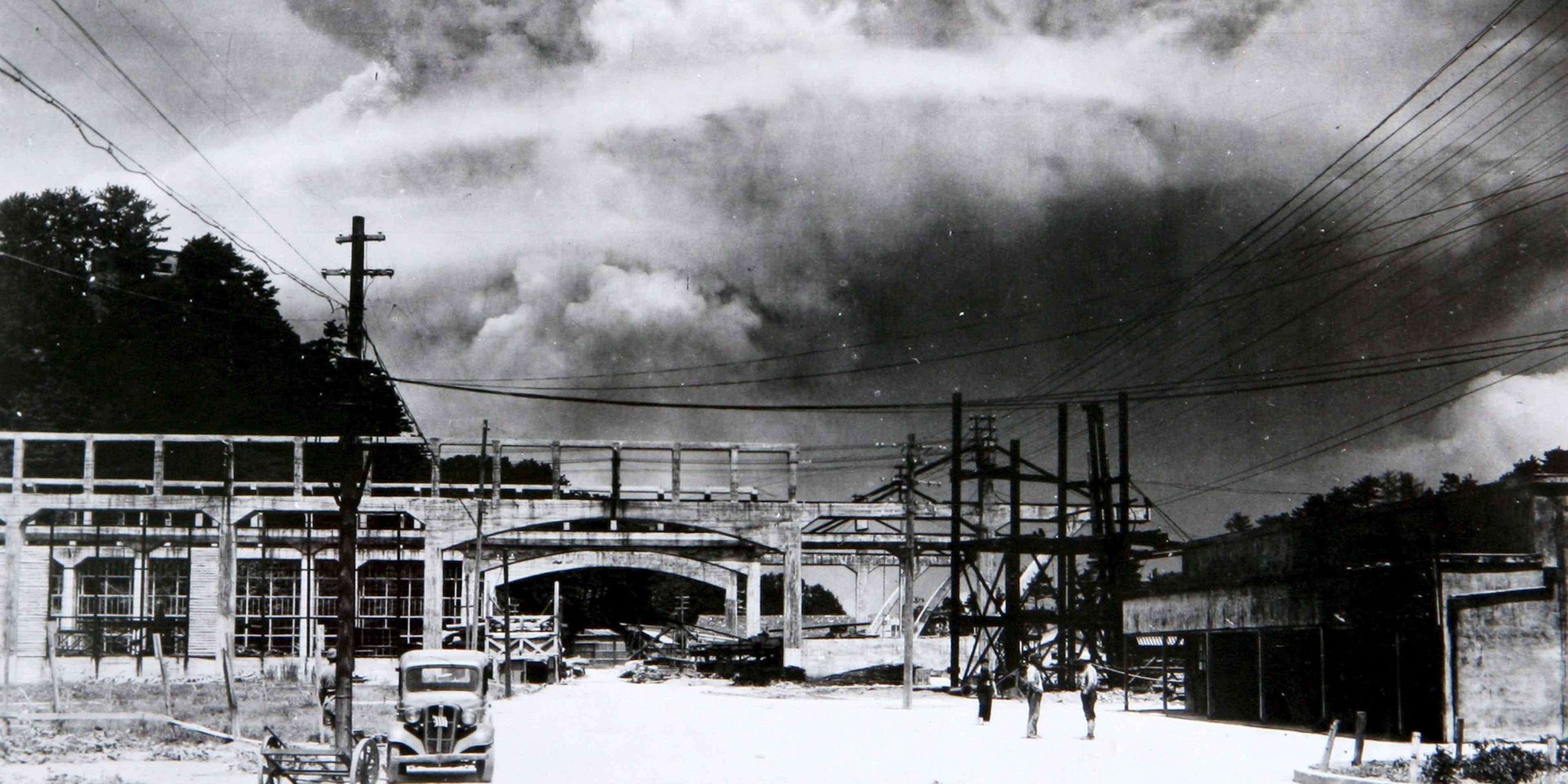 Giant mushroom cloud seen over the city of Nagasaki, with a car and people in the foreground.