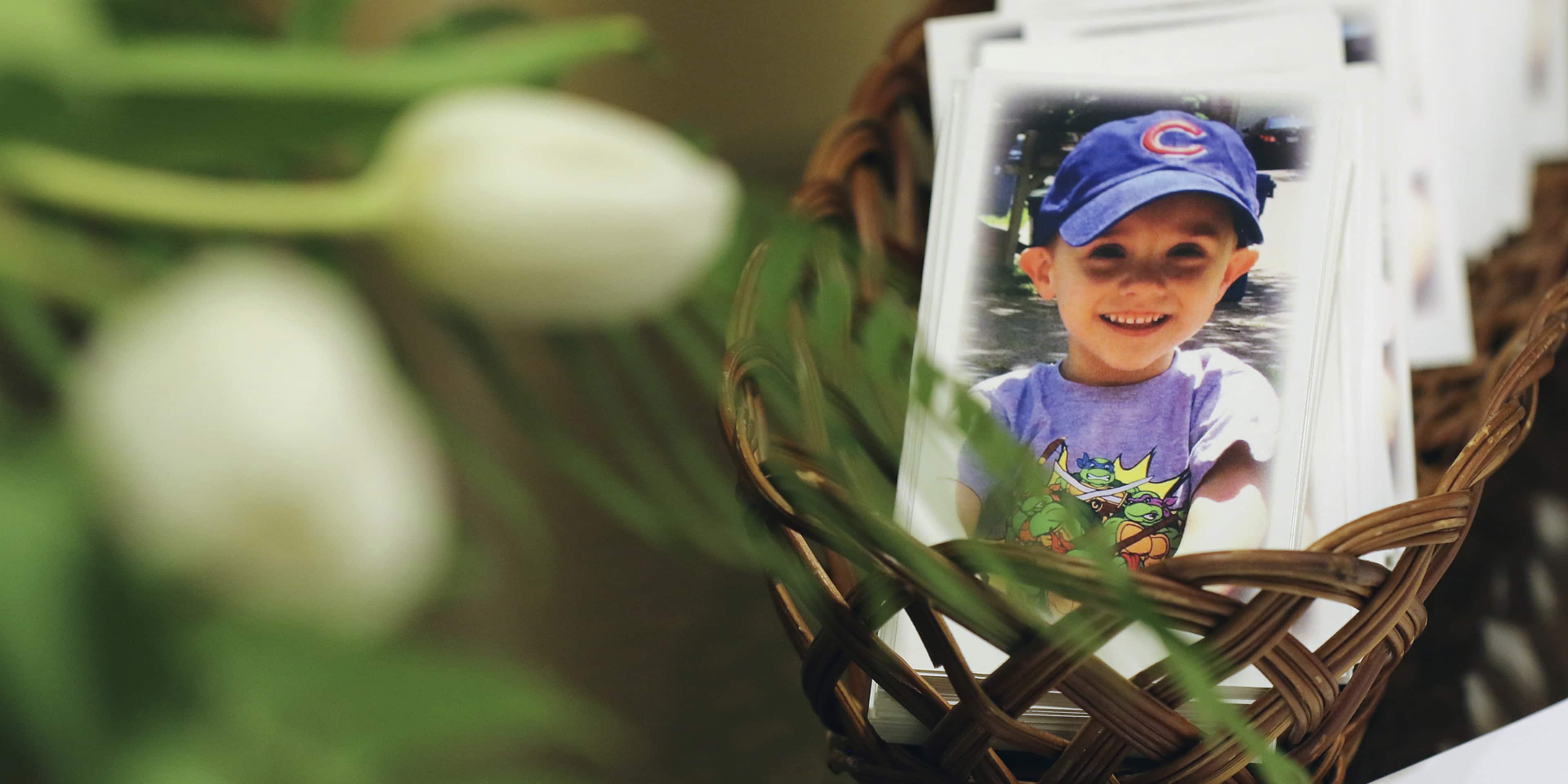 A smiling child wearing a blue cap and a purple shirt is visible through a woven basket or container, surrounded by lush greenery in the background.