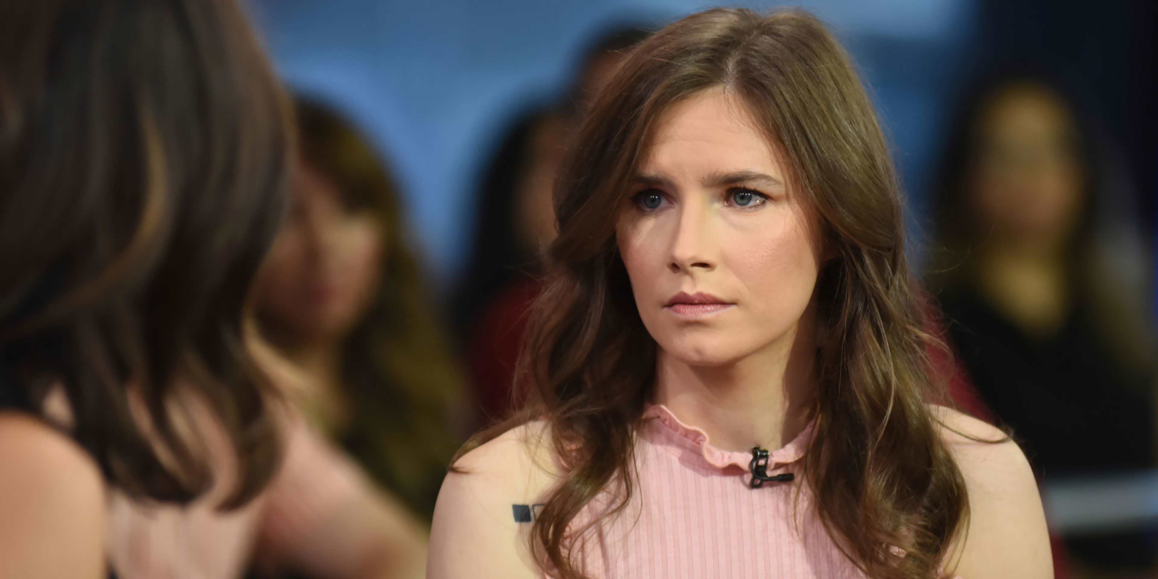 The image shows a woman with long, wavy brown hair wearing a pink top, appearing to be in a television studio or interview setting with a blurred background.
