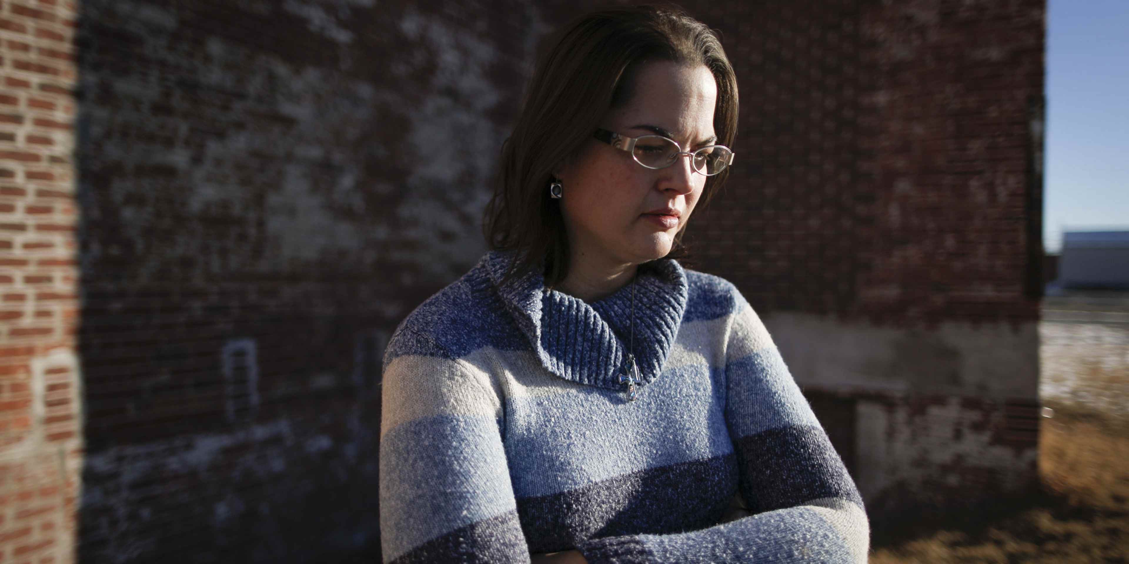 A young woman wearing a blue and gray sweater stands in front of a brick wall, her expression thoughtful as she gazes off to the side.