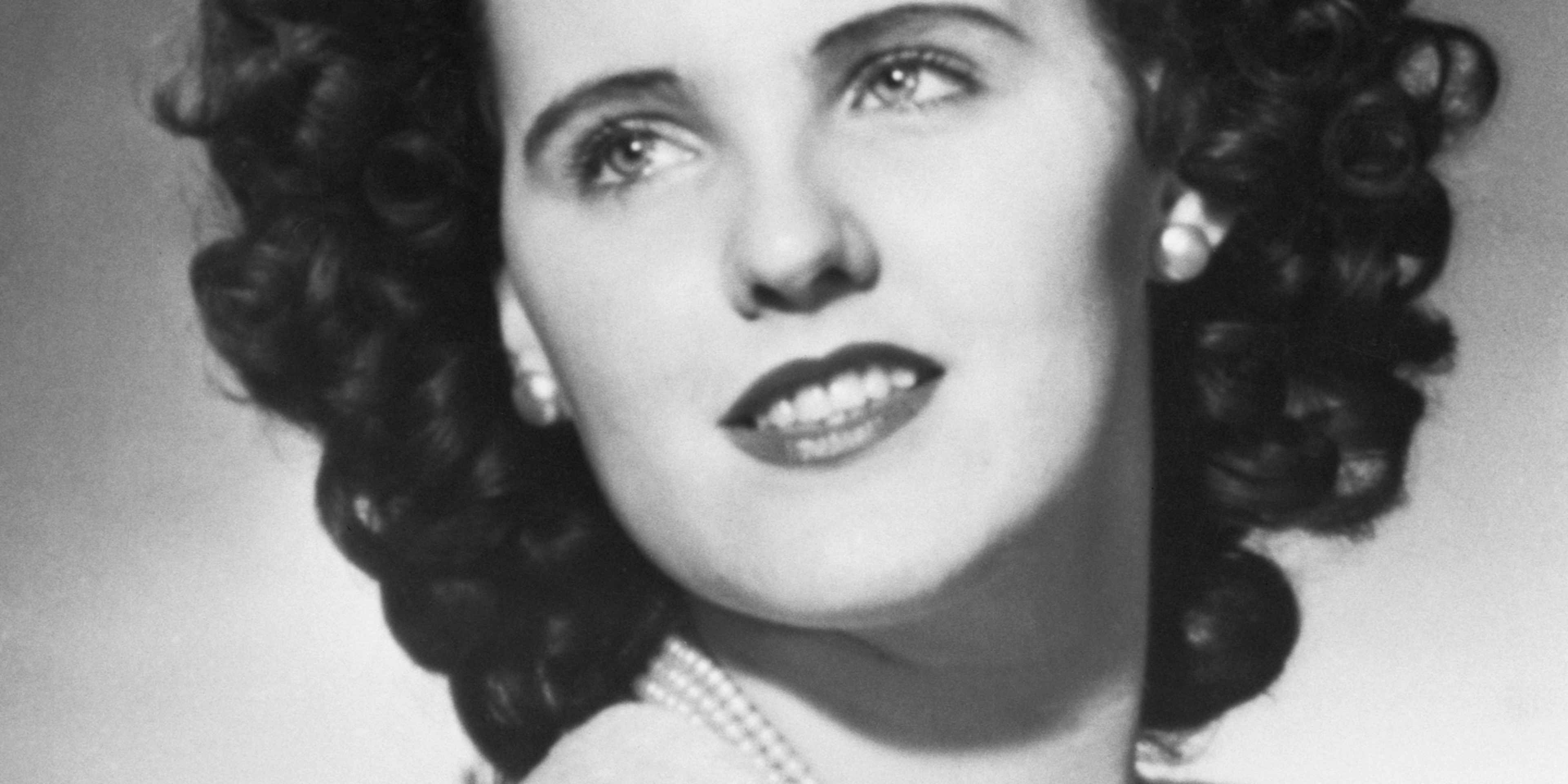 A black and white portrait of a woman with curly hair and an expressive facial expression, set against a plain background.