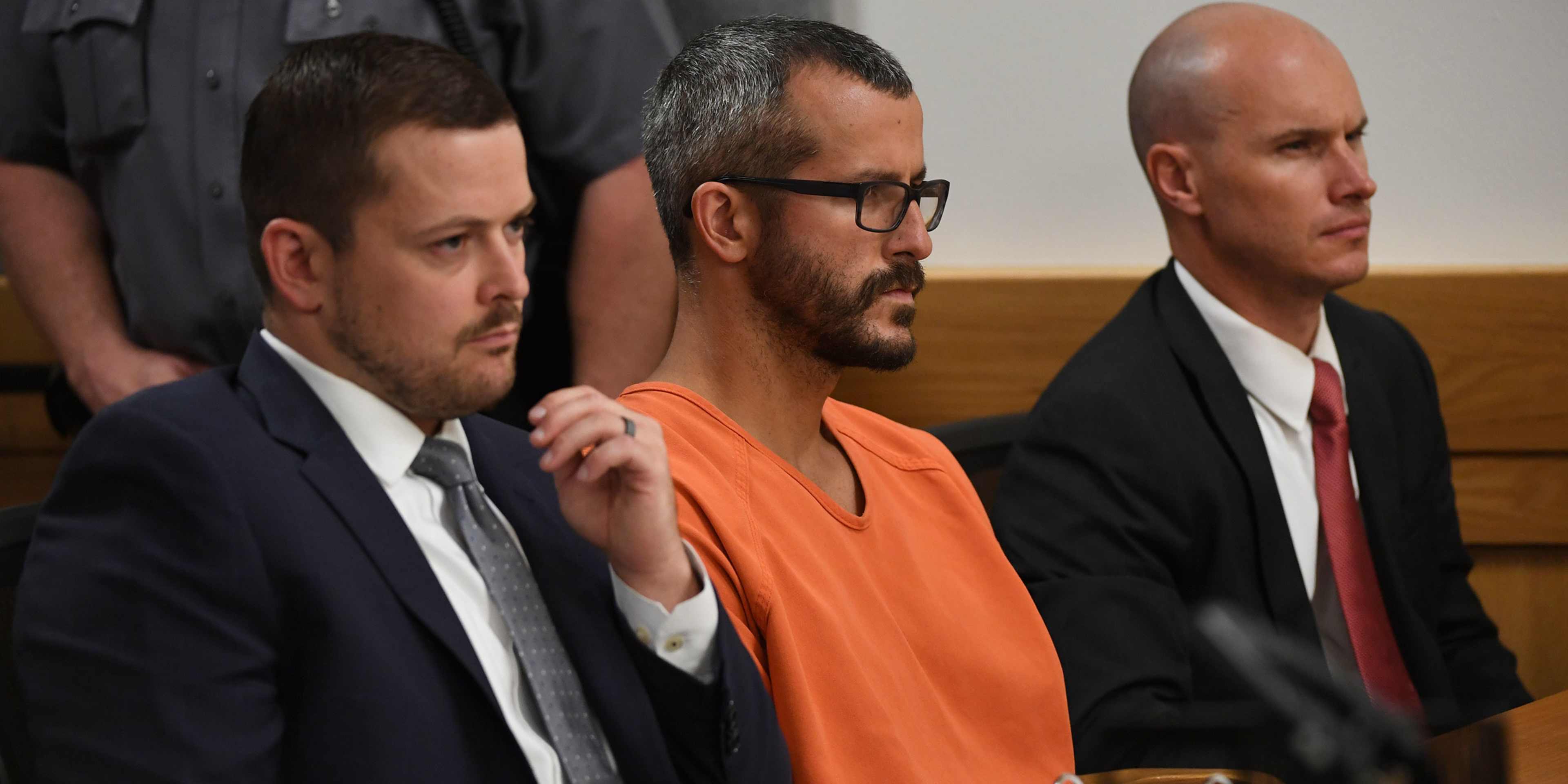 The image shows a man in an orange jumpsuit standing between two men in suits, likely in a courtroom setting.