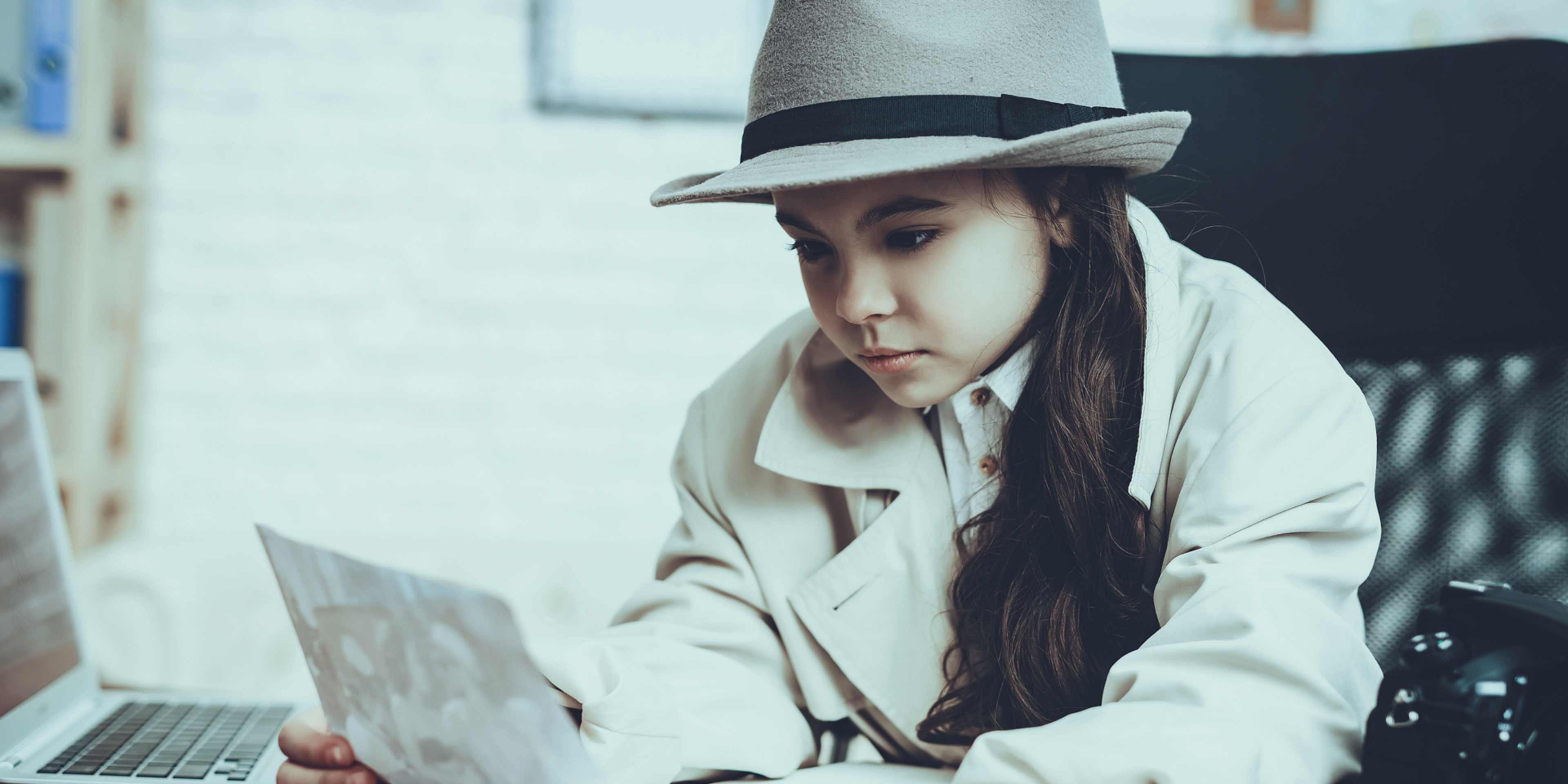 A young woman wearing a white coat and a wide-brimmed hat is sitting on a bench, intently focused on a laptop computer in front of her.