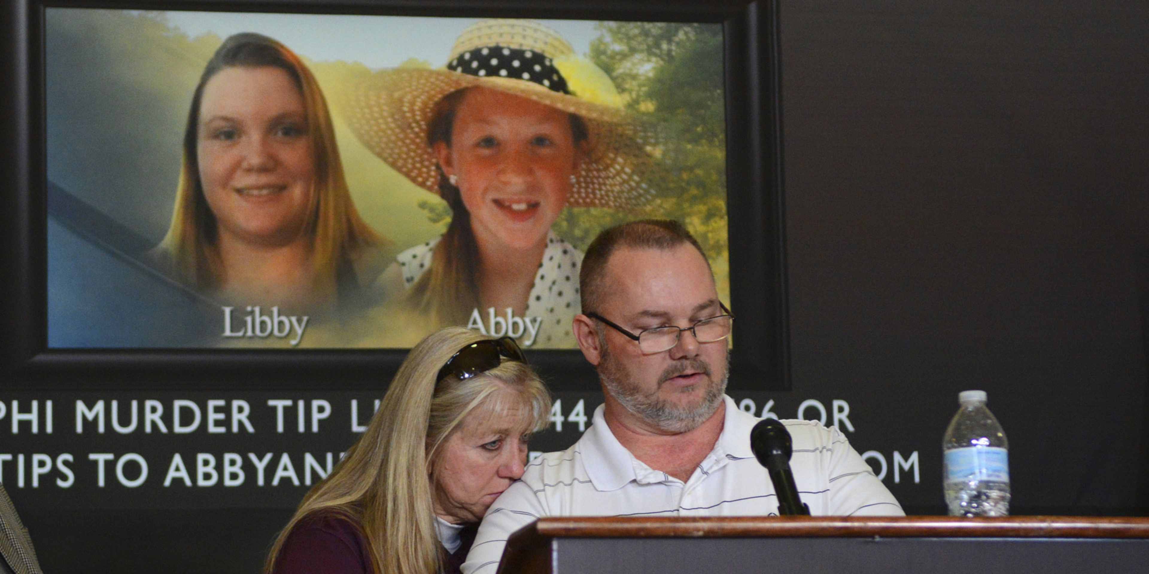 The image shows a man and a woman speaking at a podium, with a large screen behind them displaying a photograph of two smiling women.