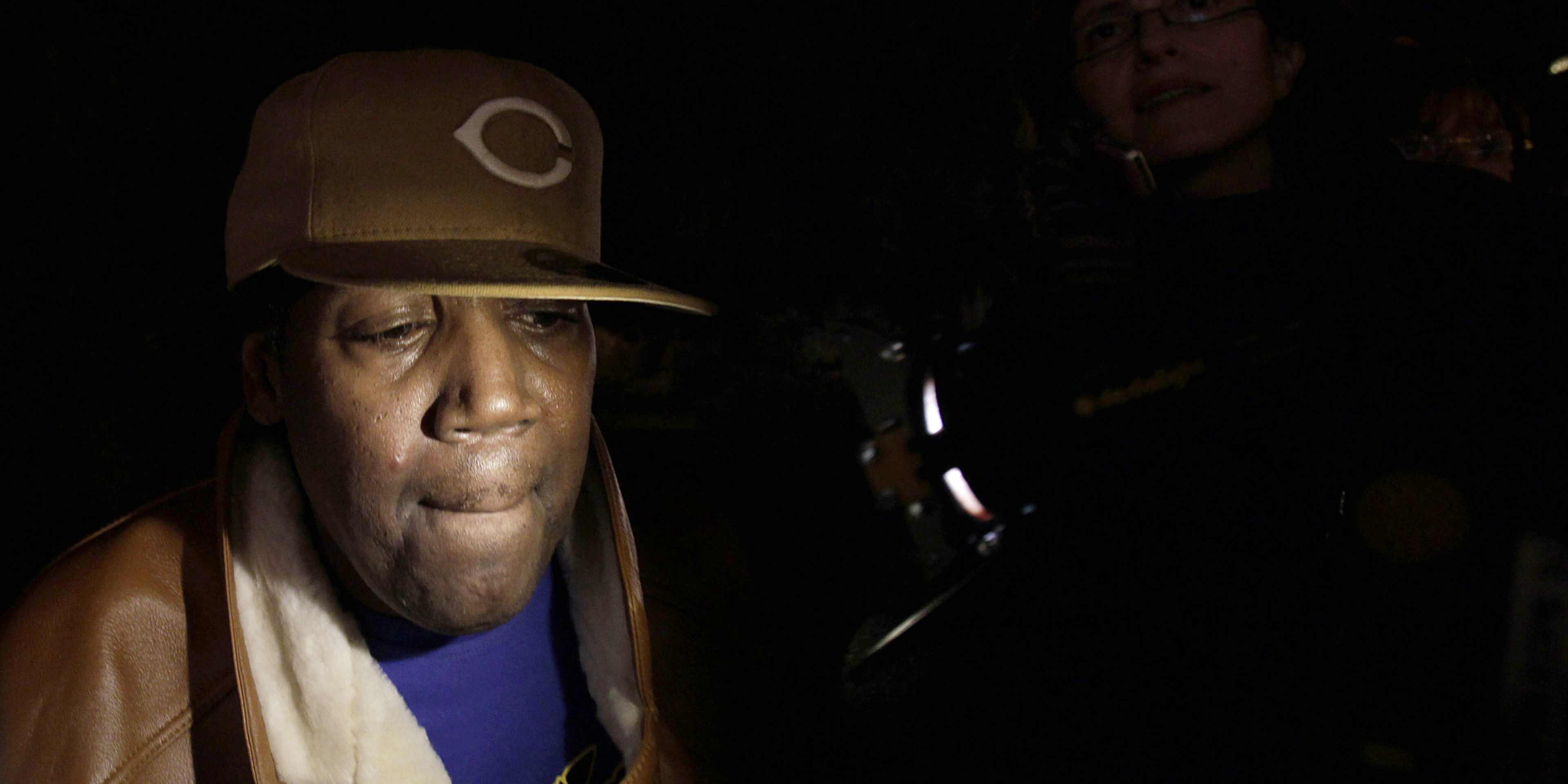 A person wearing a hard hat and a blue shirt stands in a dimly lit environment, their face partially obscured by the shadows.