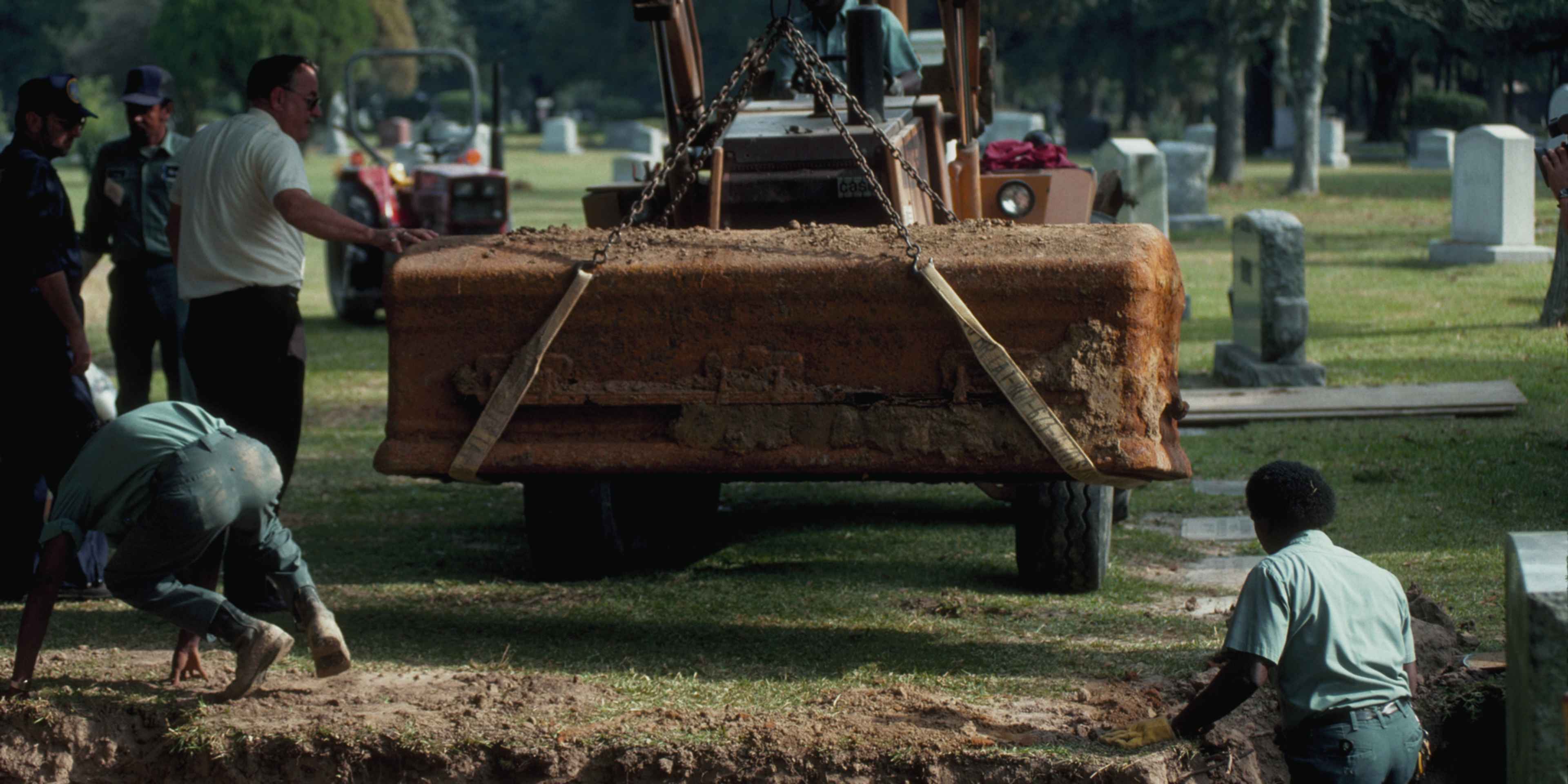 A construction site in a grassy area, with a large excavator and several workers in the foreground, and a cemetery or graveyard visible in the background.