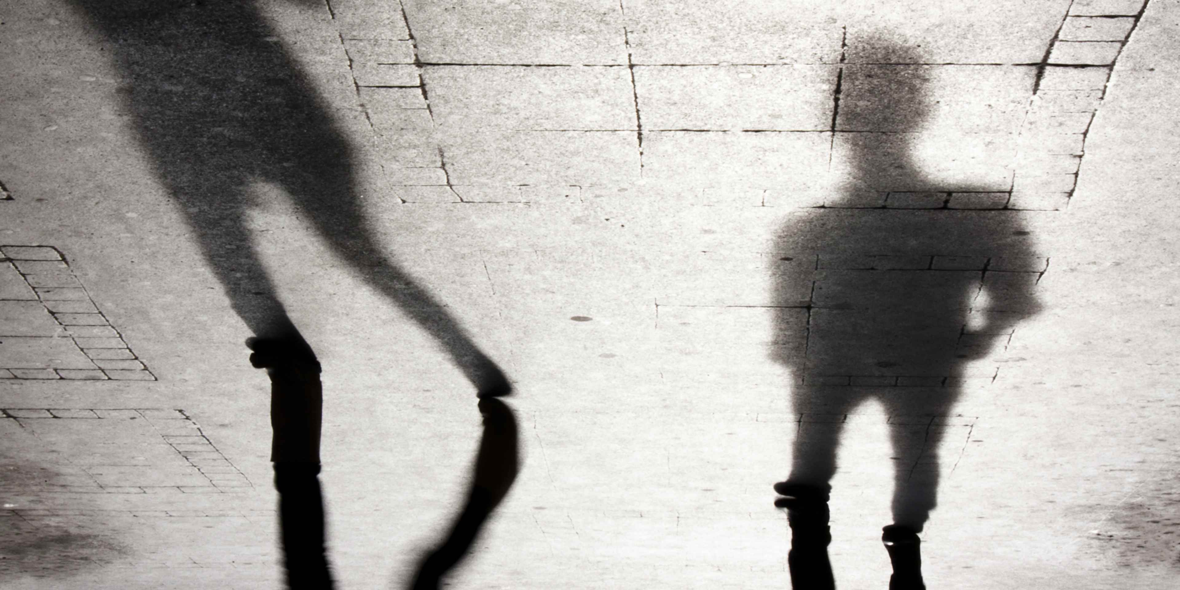 The image depicts a shadowy, silhouetted figure standing in a dimly lit, abstract background with geometric shapes and patterns.