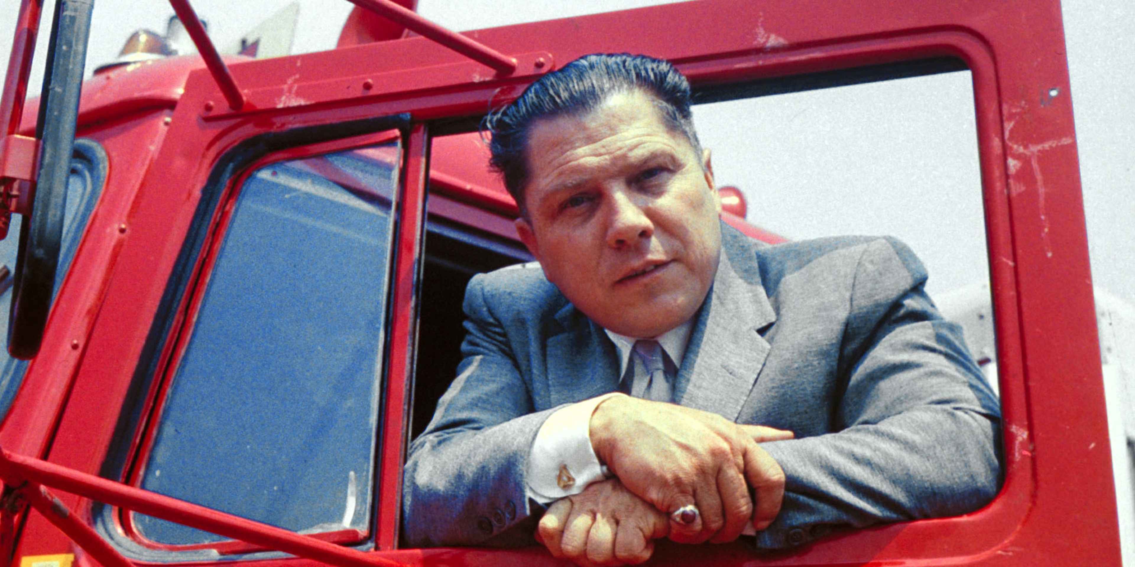 A man in a suit is sitting in the driver's seat of a red truck, looking out the window.