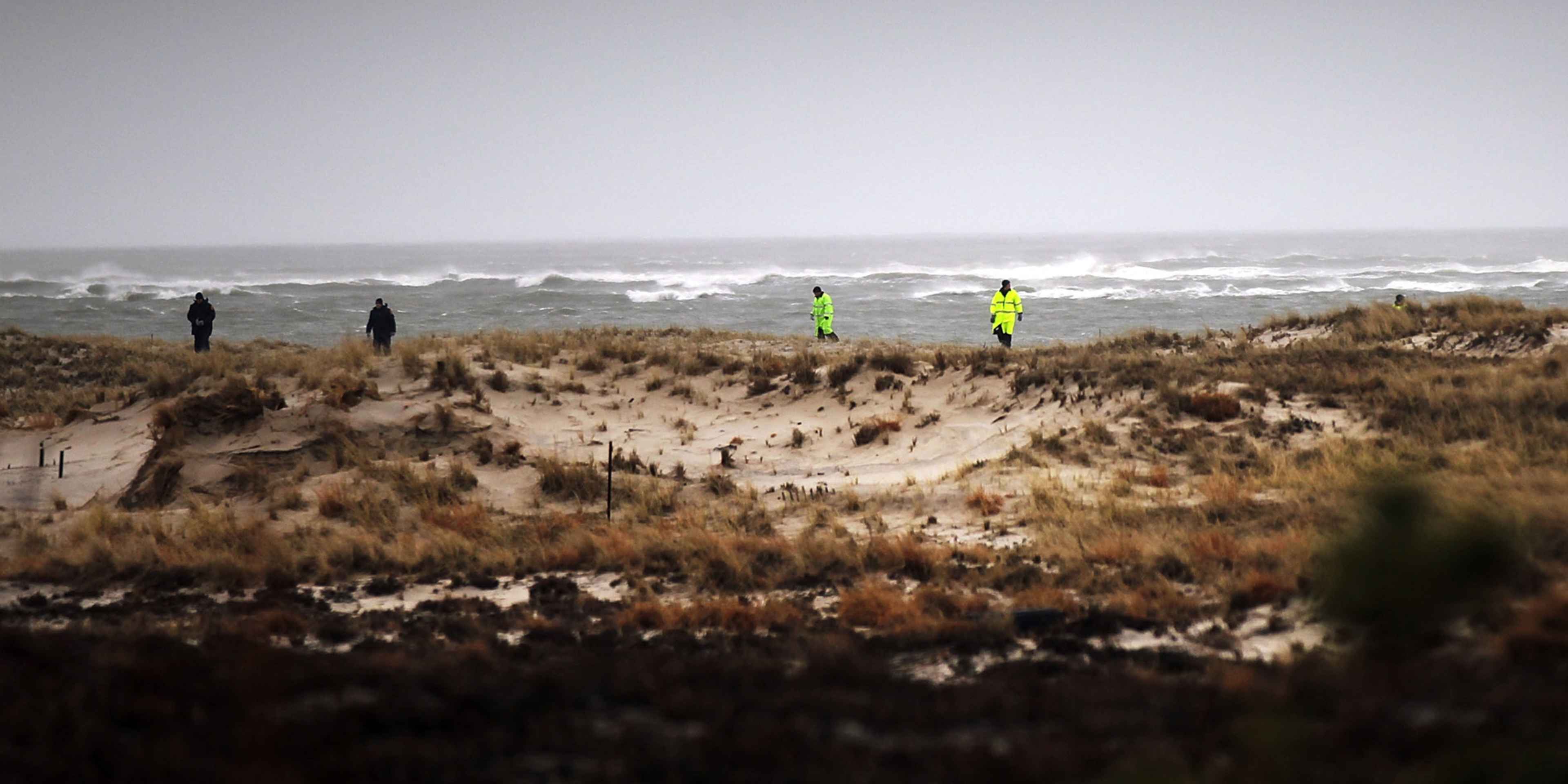 The image depicts a rugged, windswept coastal landscape with two people in the distance, wearing bright yellow safety vests, walking along a path through the dunes and grassy vegetation.
