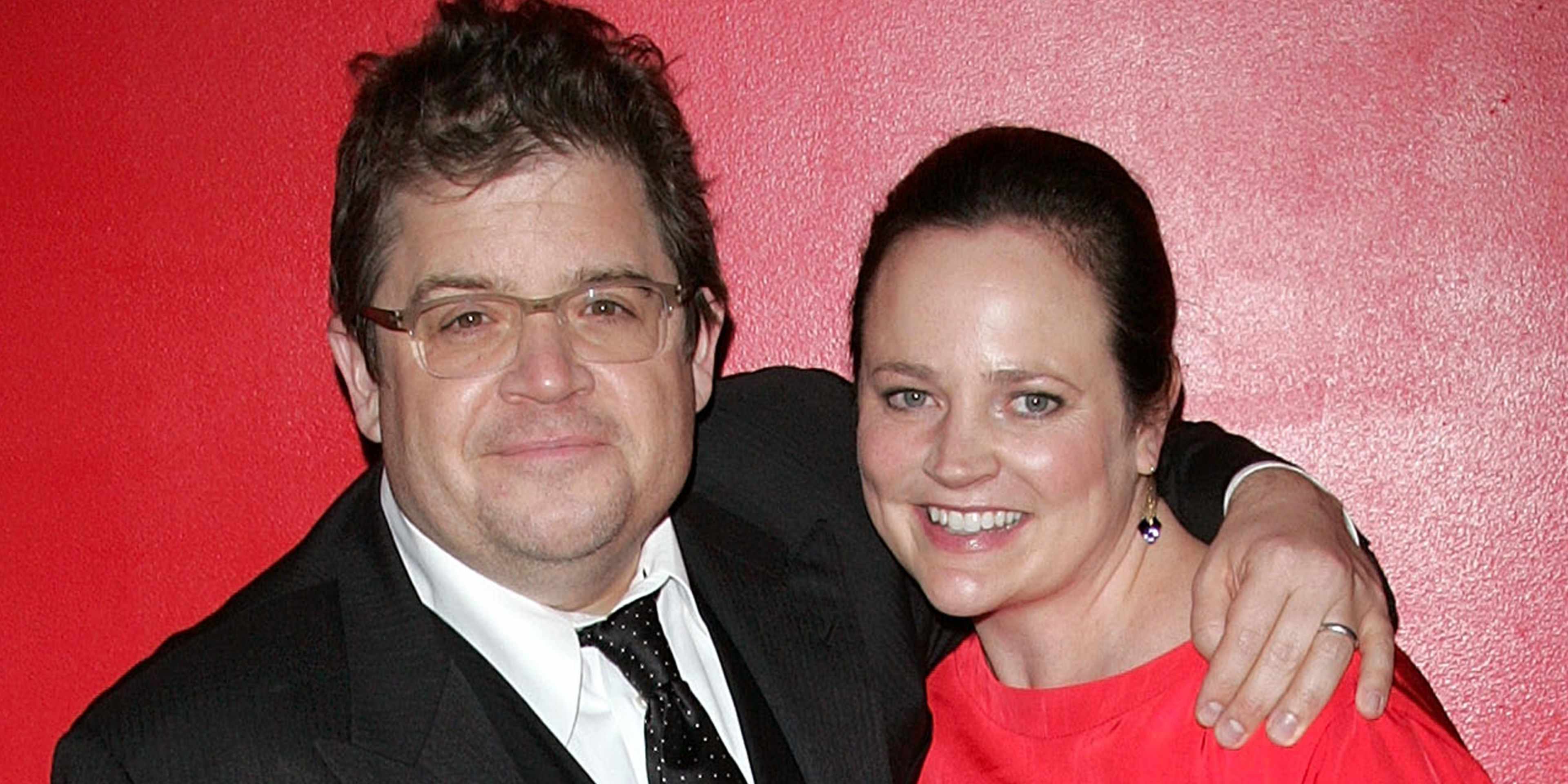 The image shows a middle-aged man and woman posing together in front of a vibrant red background, both dressed formally and smiling at the camera.