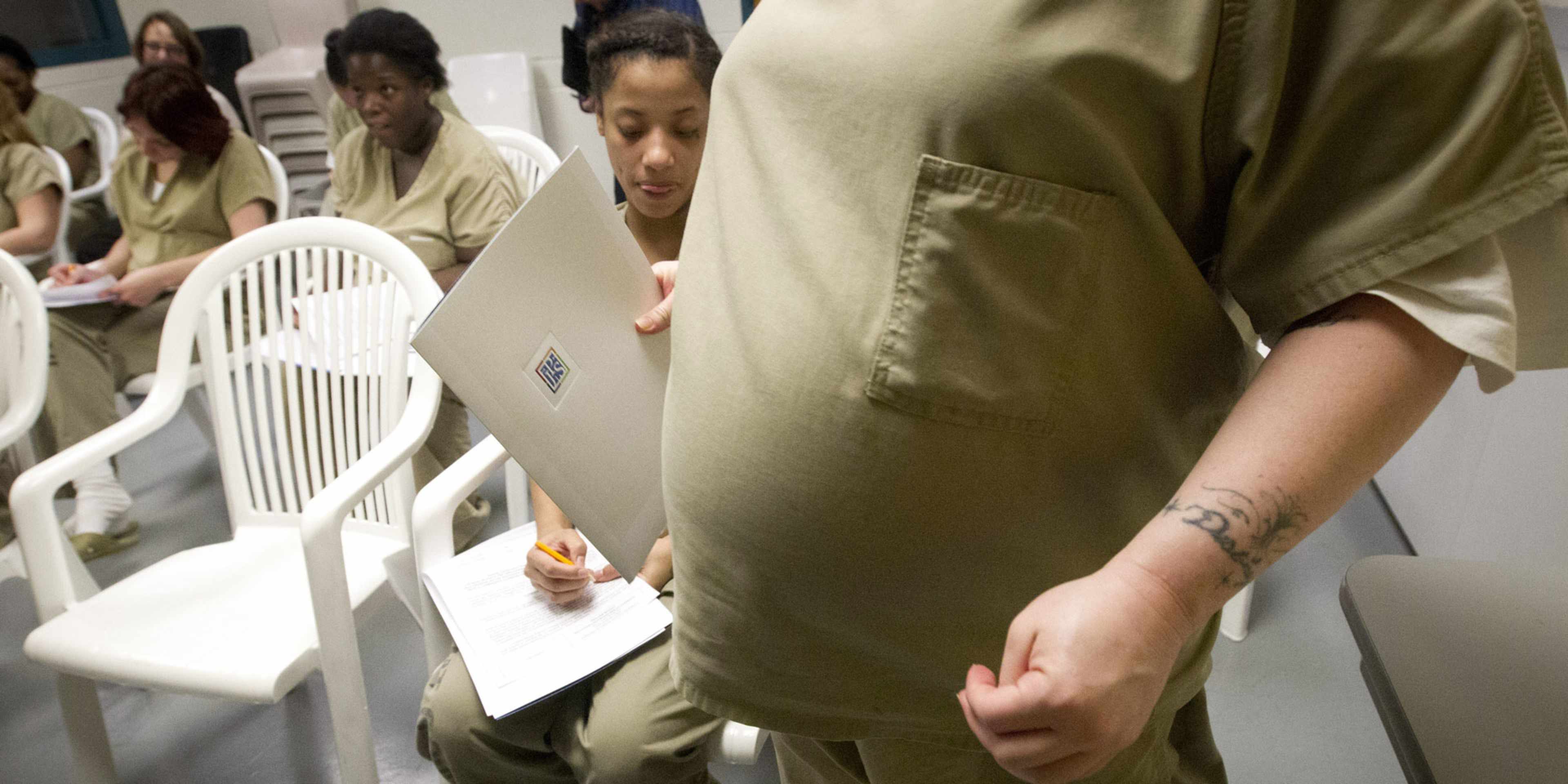 The image shows a group of people, likely students or trainees, seated at desks in what appears to be a classroom or study area. They are all wearing similar olive-colored uniforms, and one person in the foreground is holding a document or paper.