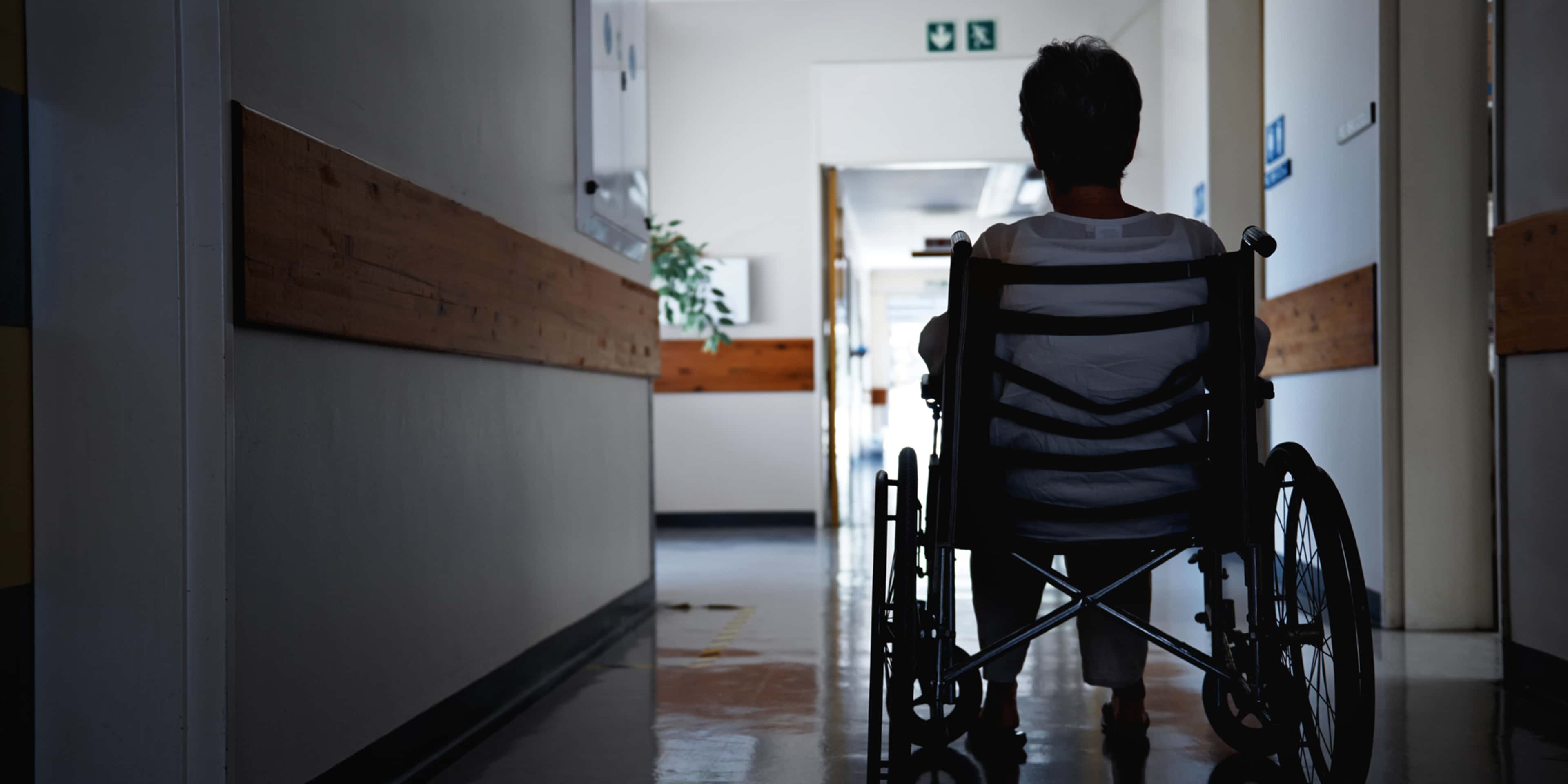A person in a wheelchair is seen moving down a hospital corridor, with a door and other details visible in the background.