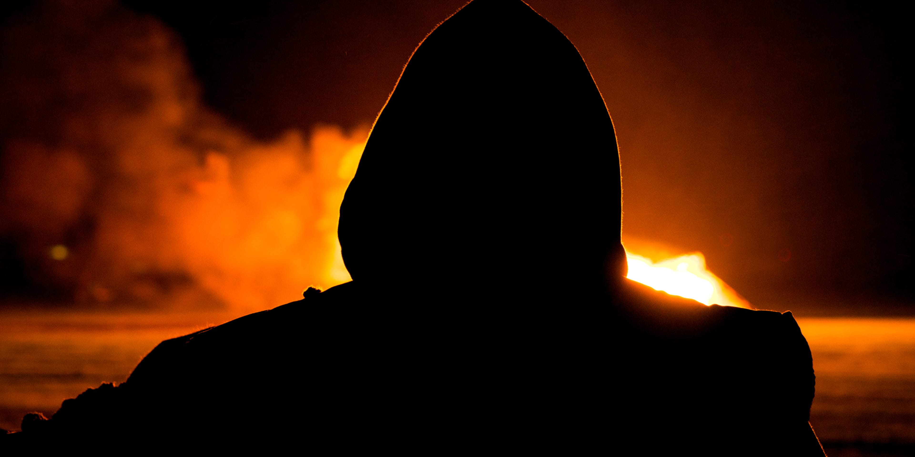 A hooded figure stands silhouetted against a fiery, orange-hued background, creating an ominous and foreboding atmosphere.