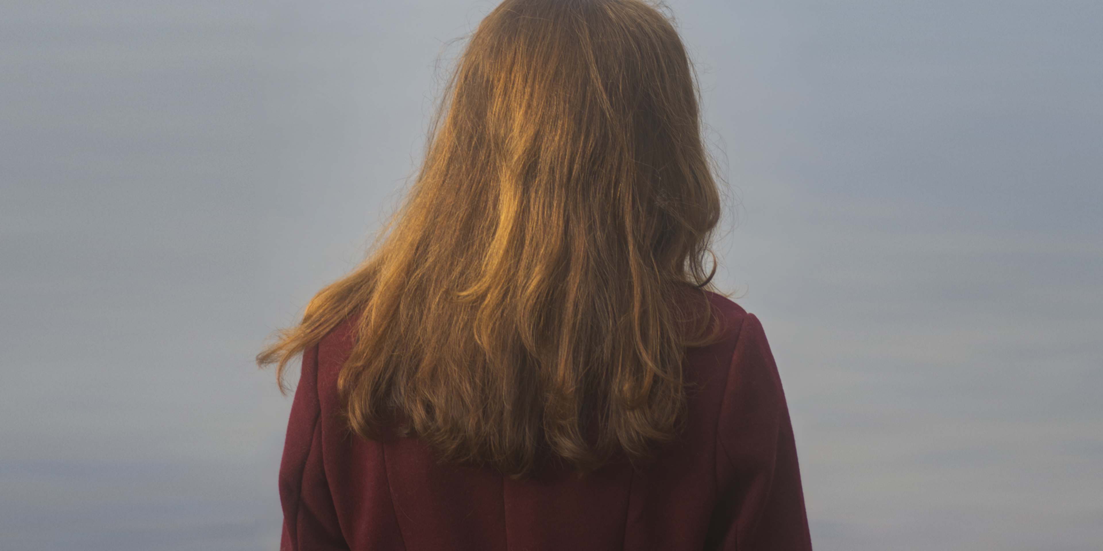 A person with long, flowing brown hair wearing a burgundy-colored garment stands facing away from the camera, with a cloudy sky visible in the background.