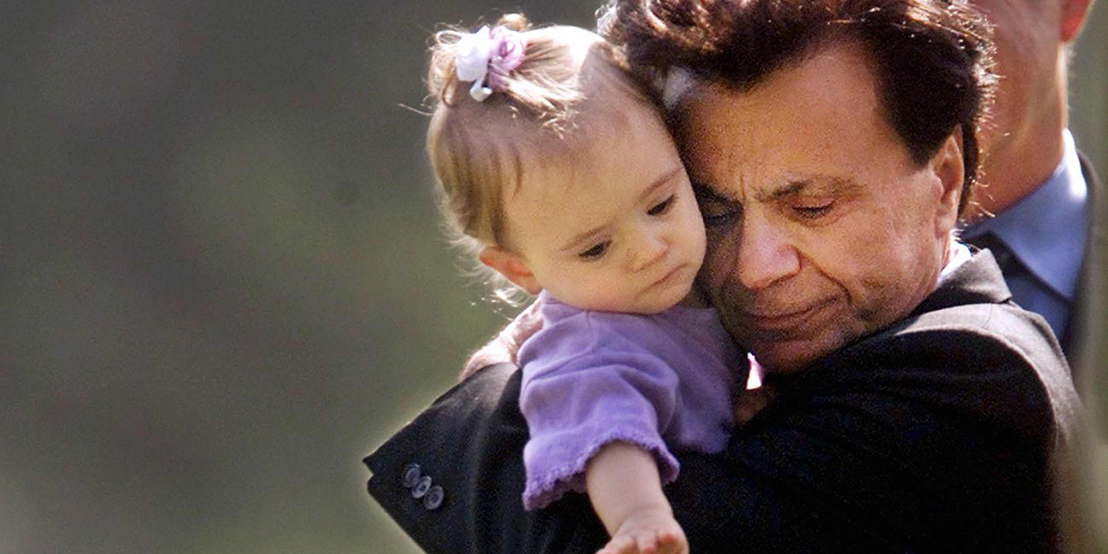 A man in a dark suit is holding a young girl in a purple dress, with both of them appearing to have a tender, affectionate interaction.