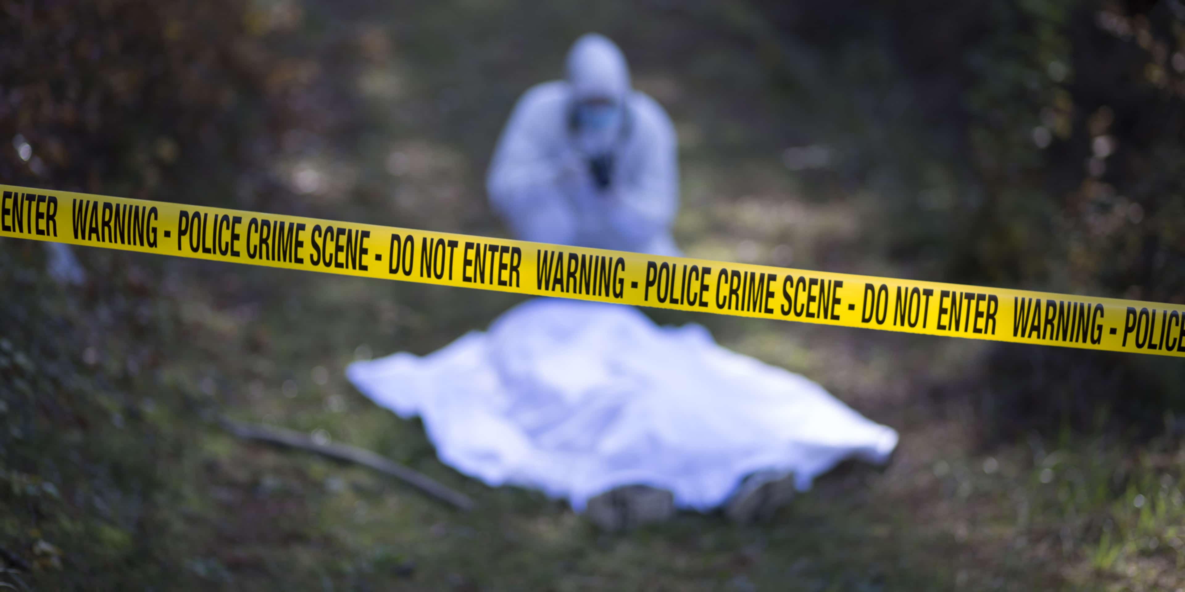 The image shows a white sheet or cloth covered by yellow police tape, with a dark, blurred background suggesting a wooded or natural setting.