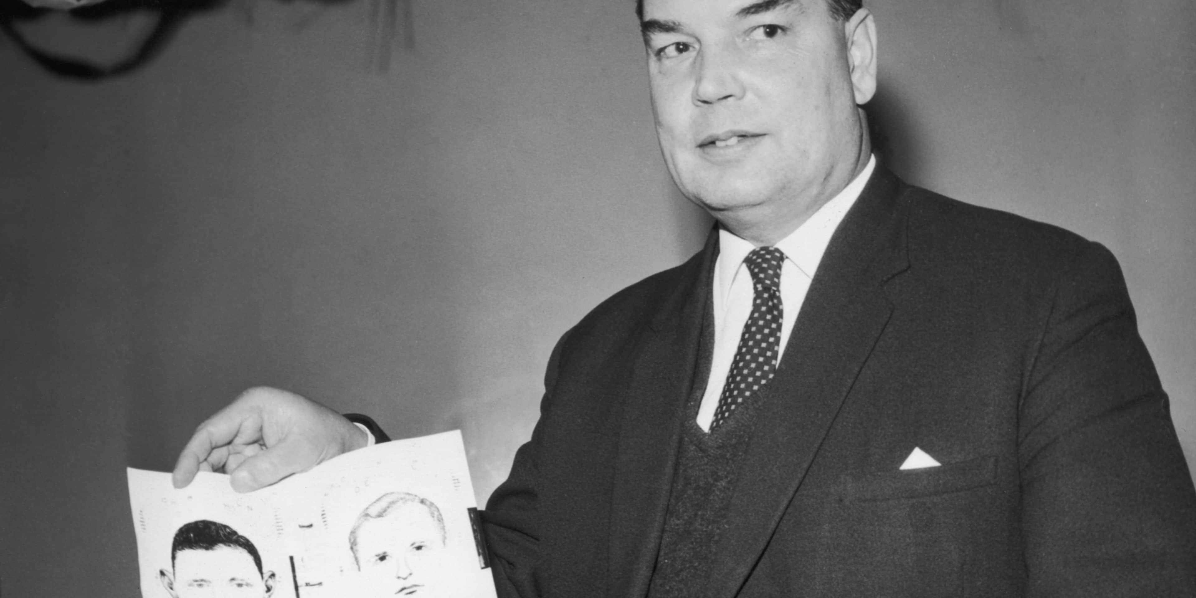 A middle-aged man in a suit holding a piece of paper with cartoon-like drawings on it, standing in front of a plain background.