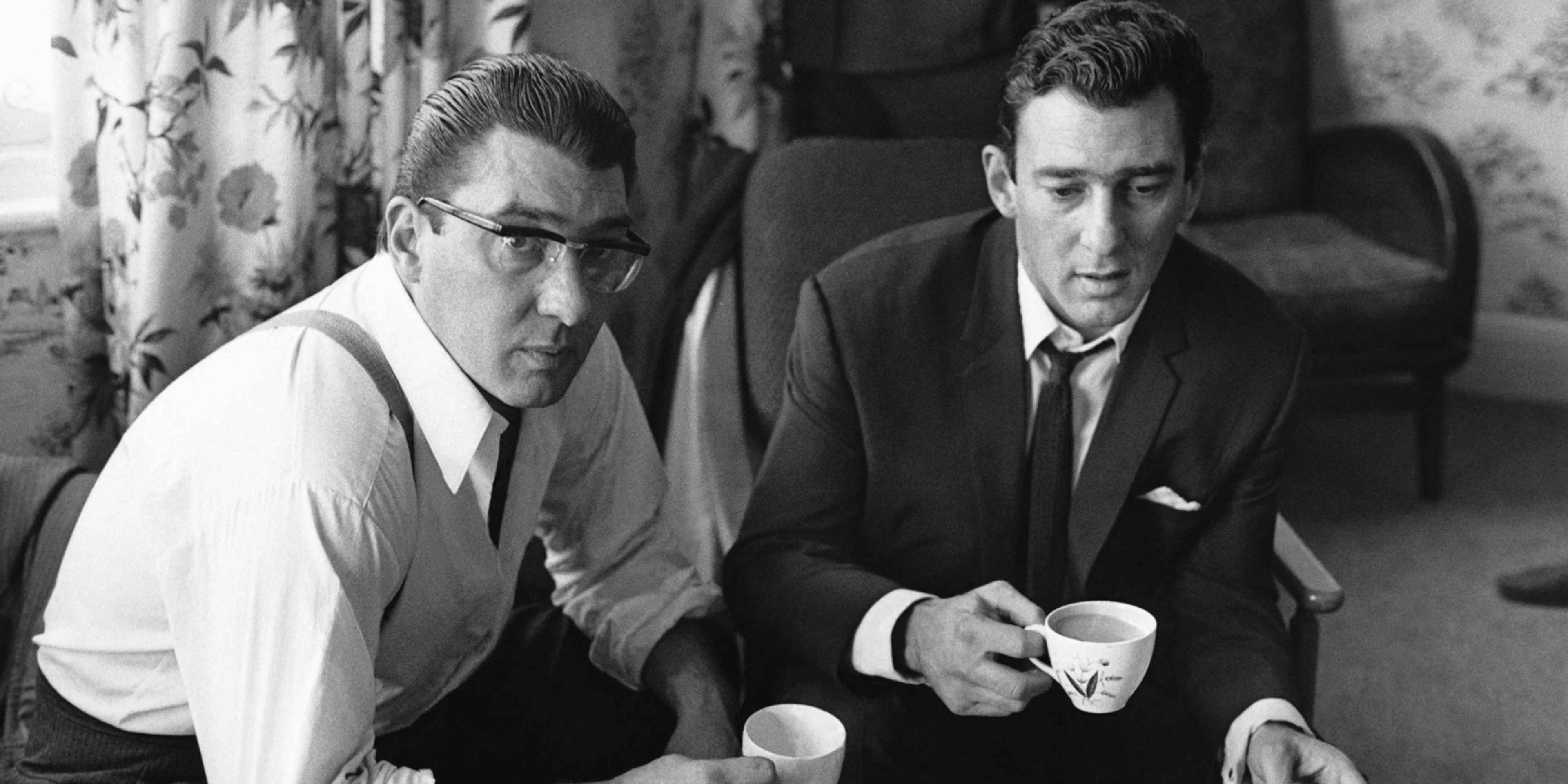 Two men in suits sitting together, with a floral curtain in the background.