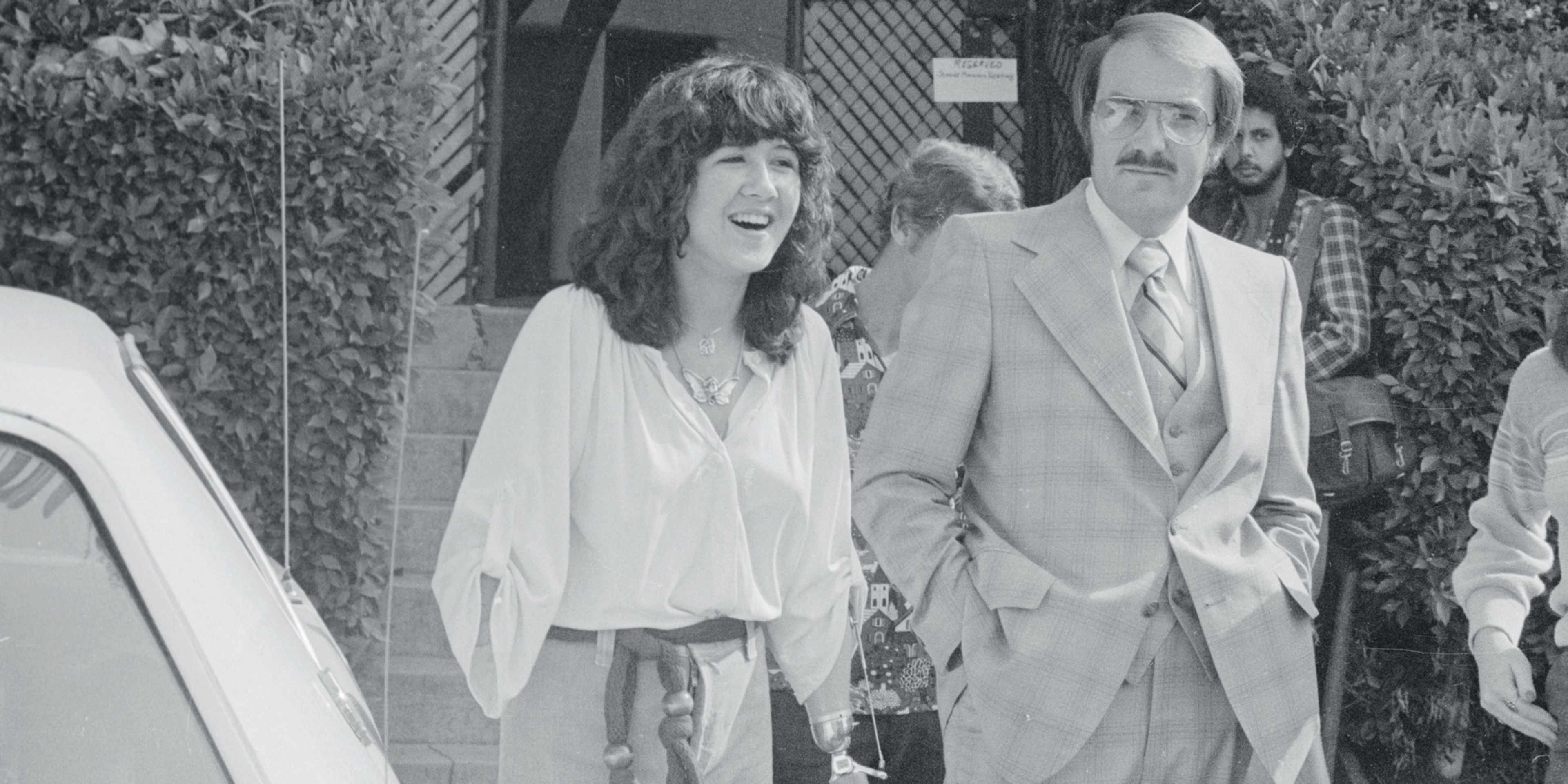 A man and a woman, both dressed formally, are walking together in an outdoor setting with a fence and foliage visible in the background.