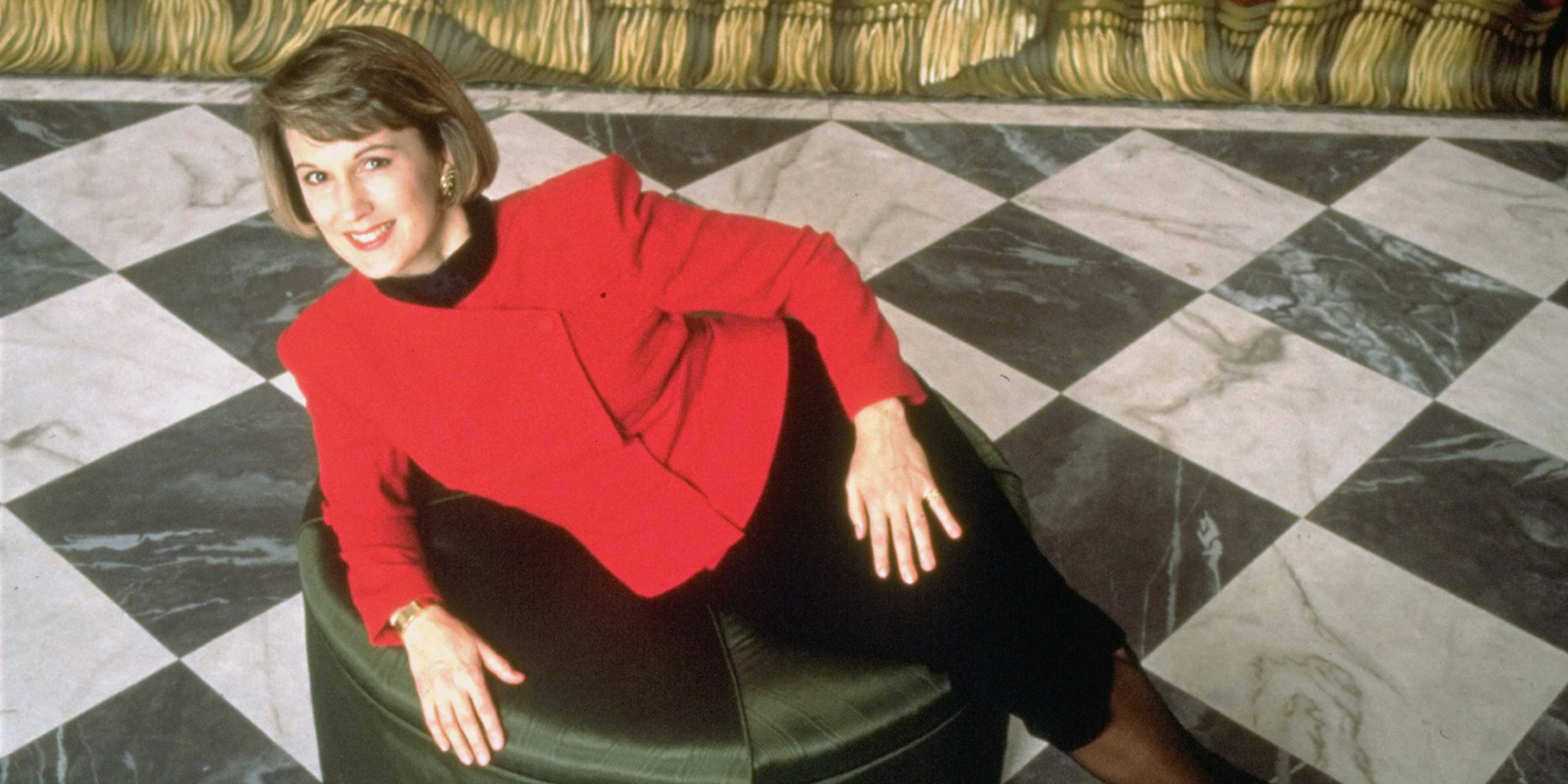 A woman in a red sweater stands on a checkered floor against a backdrop of curtains.