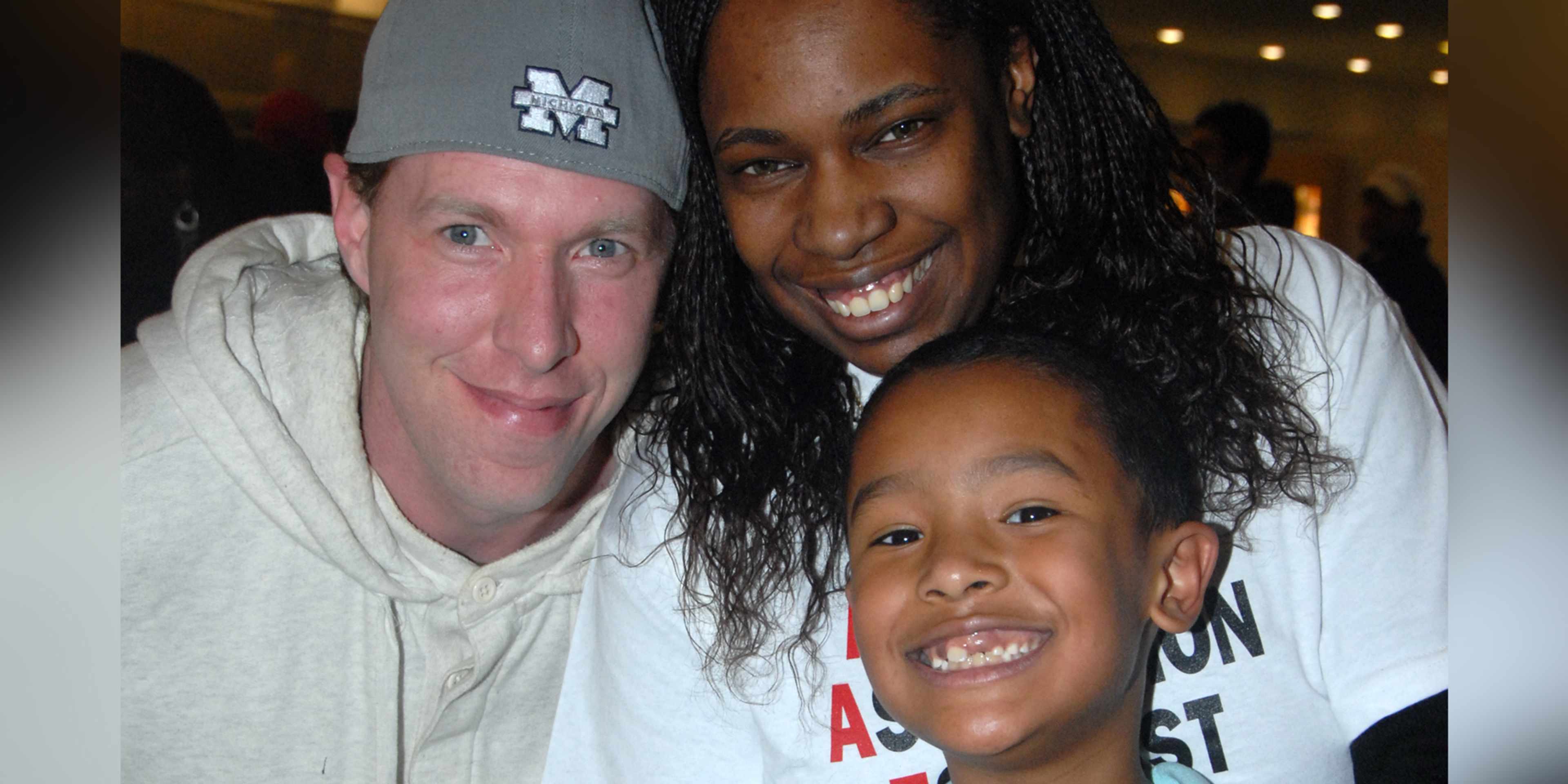 The image shows a smiling group of three people, two adults and one child, posing together in what appears to be a casual setting with some lighting fixtures visible in the background.