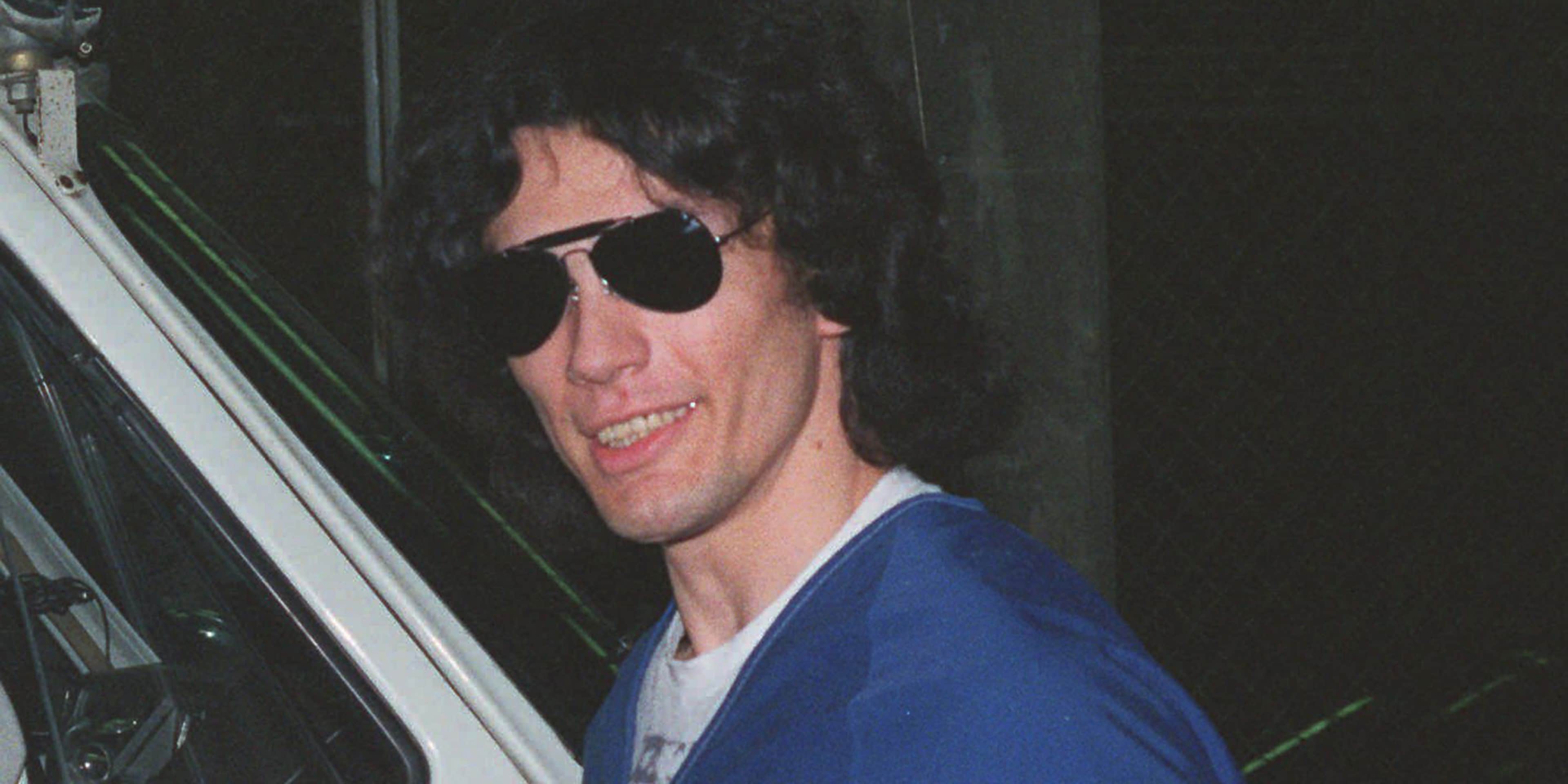 A person wearing sunglasses and a blue jacket is visible in the foreground, with a car visible in the background.