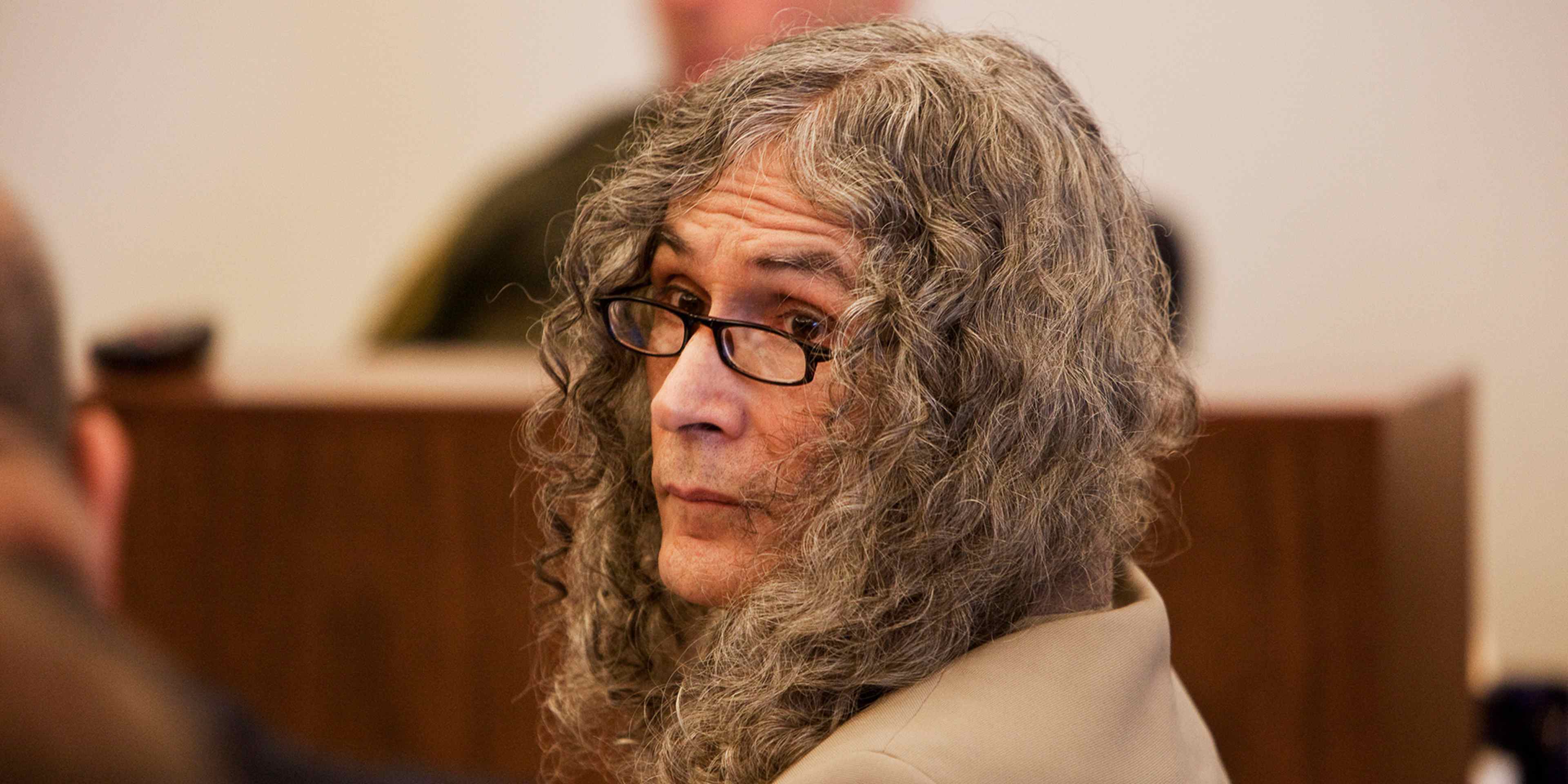 The image shows an elderly person with long, unkempt gray hair and glasses, sitting in what appears to be a courtroom or legal setting.