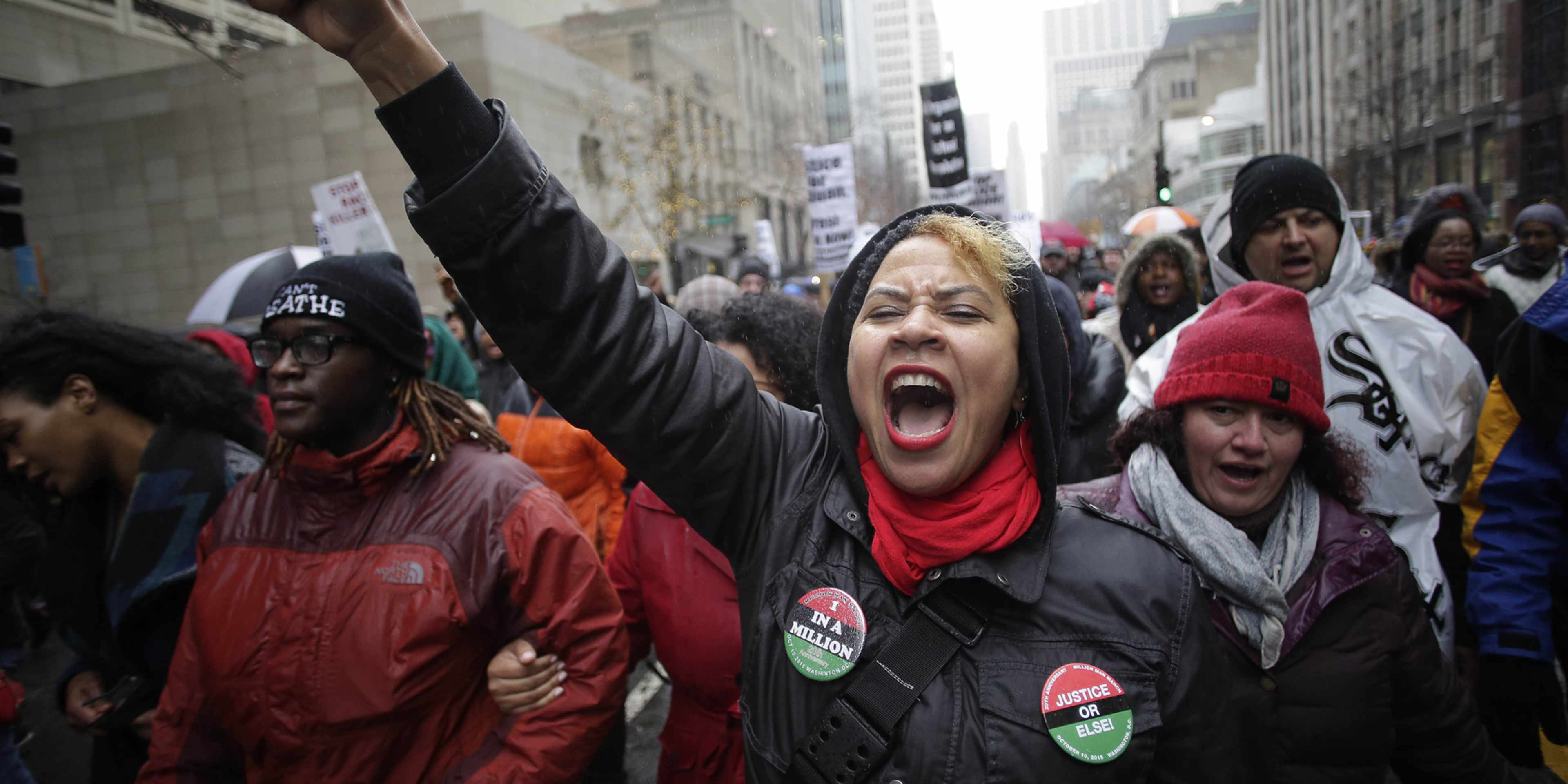 A group of people, some wearing red and black clothing, are gathered in a city street, with their arms raised and expressions of protest or activism.