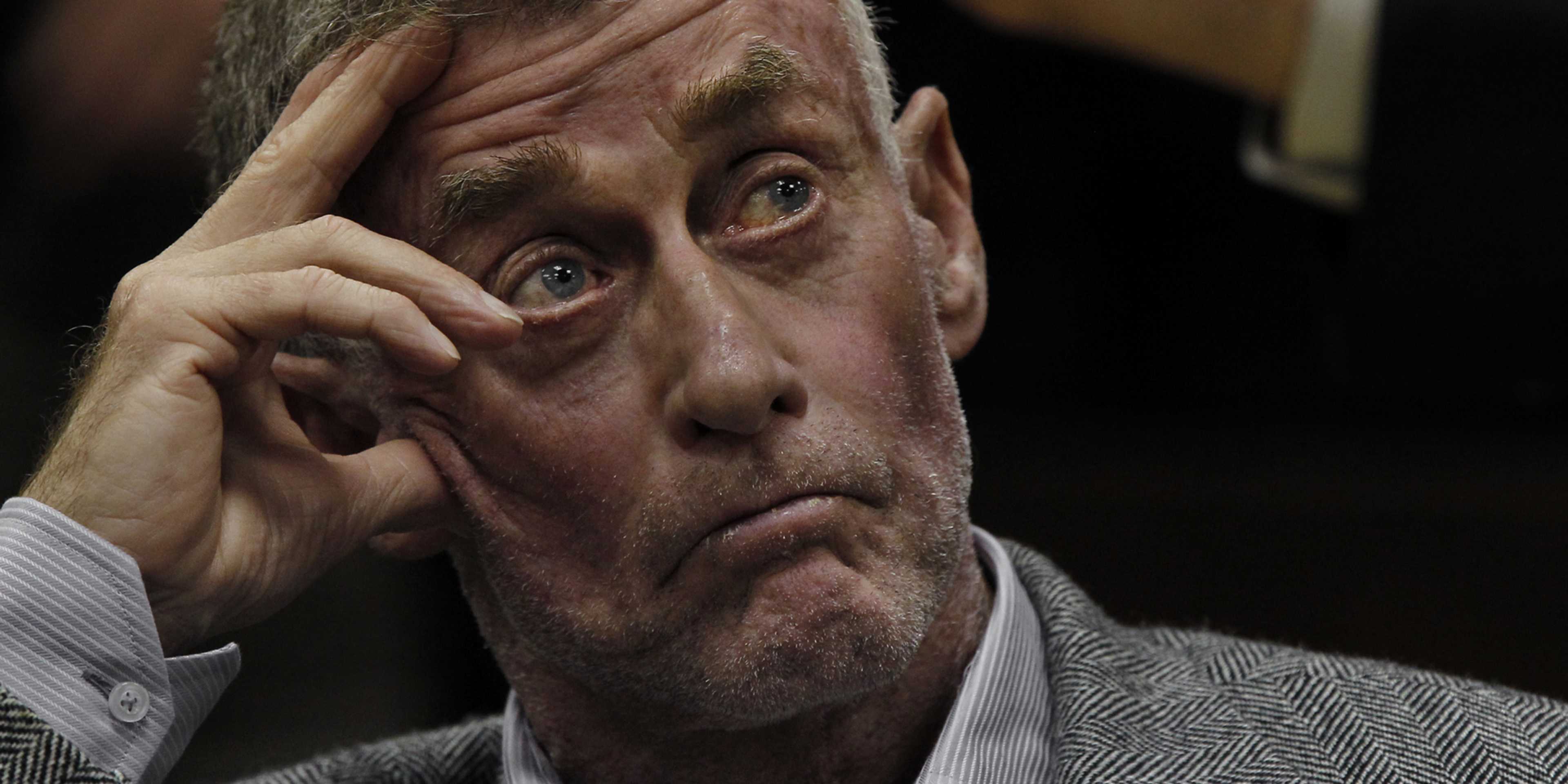 A middle-aged man with a serious expression, his hand on his forehead, appears to be deep in thought or troubled, set against a dark background.