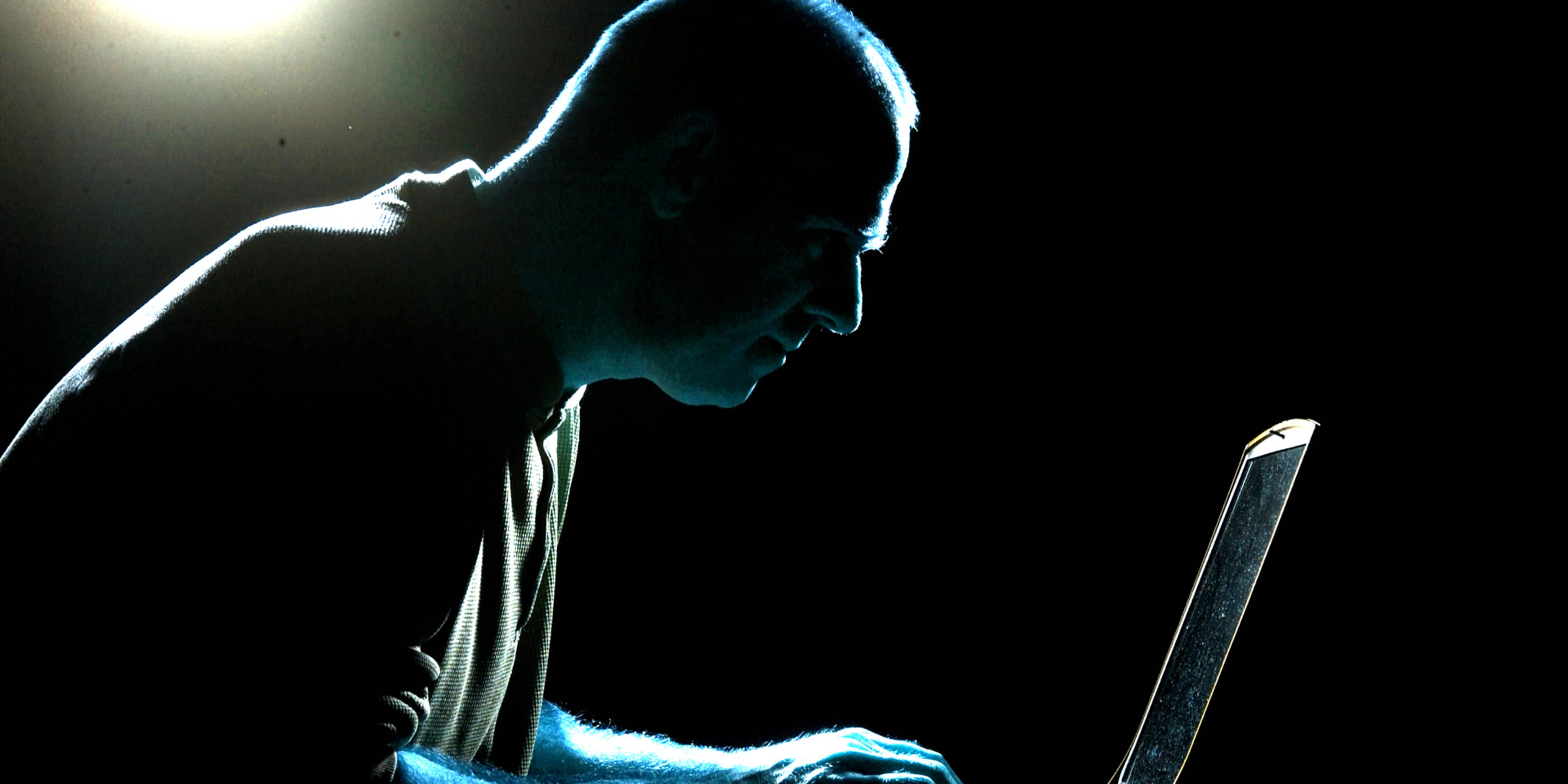 A hooded figure is intently focused on a laptop computer, casting a dramatic silhouette against the dark background.