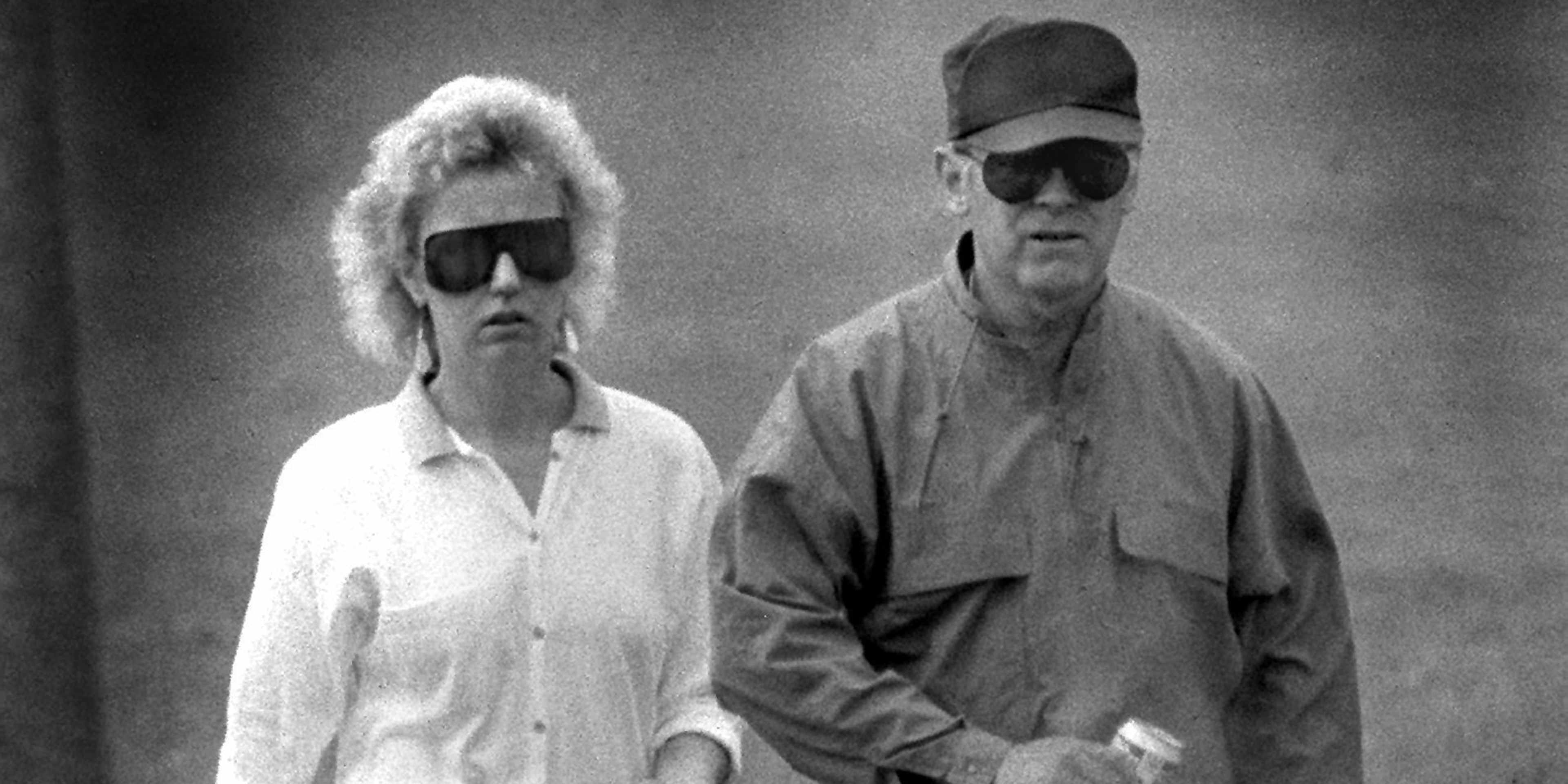 Two individuals, a man and a woman, wearing sunglasses and casual attire, standing together in a black and white photograph.