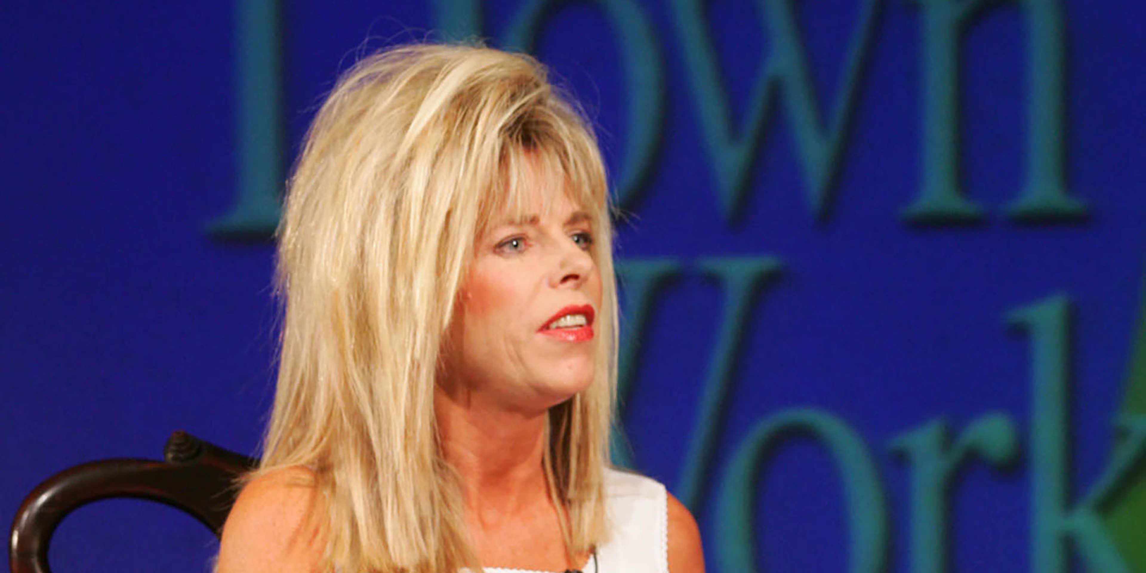 A blonde woman with a serious expression is seated in front of a vibrant blue background.