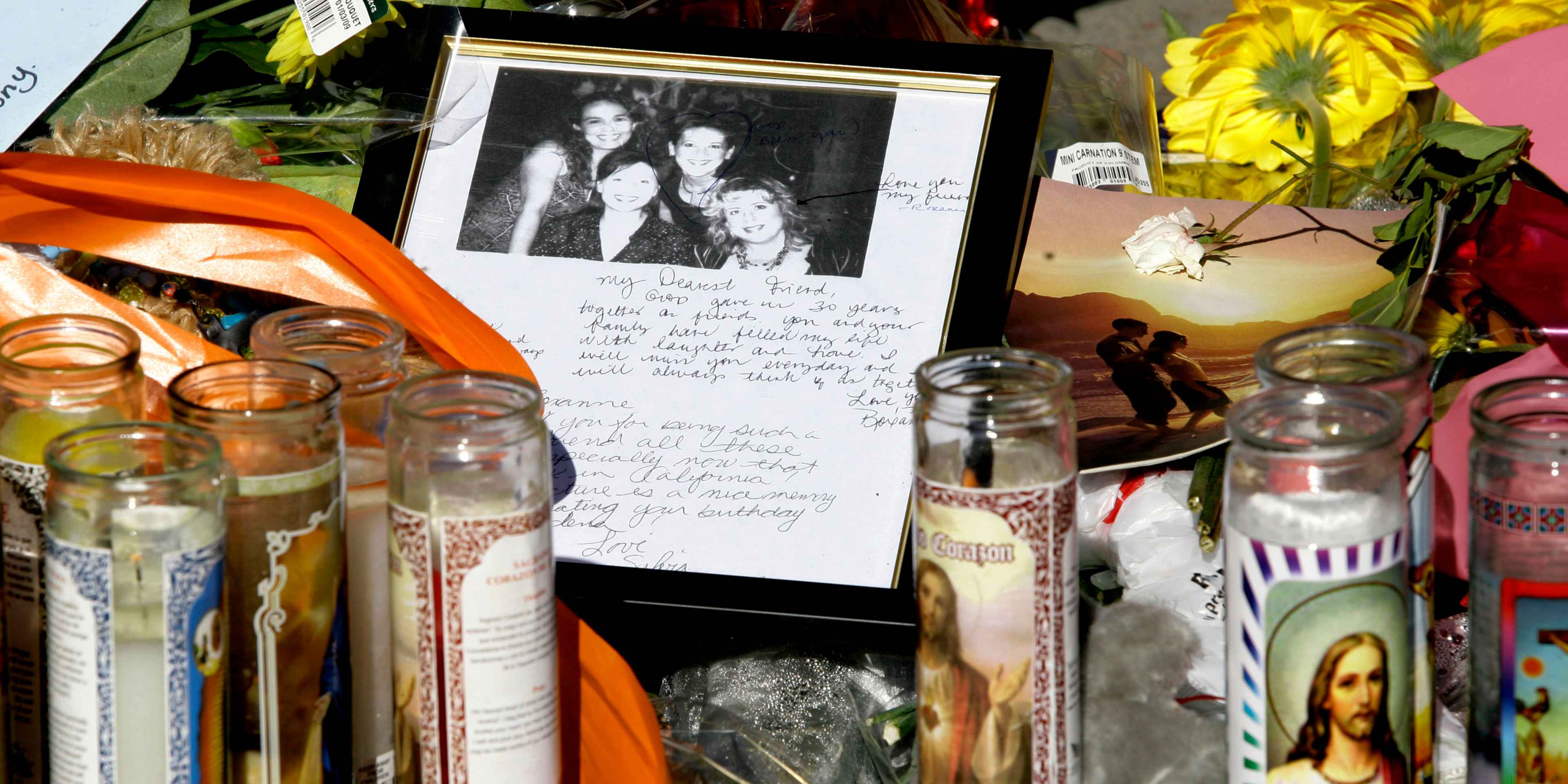The image shows a framed photograph of several people surrounded by various memorial items, including candles, flowers, and handwritten notes, against a backdrop of colorful decorations.