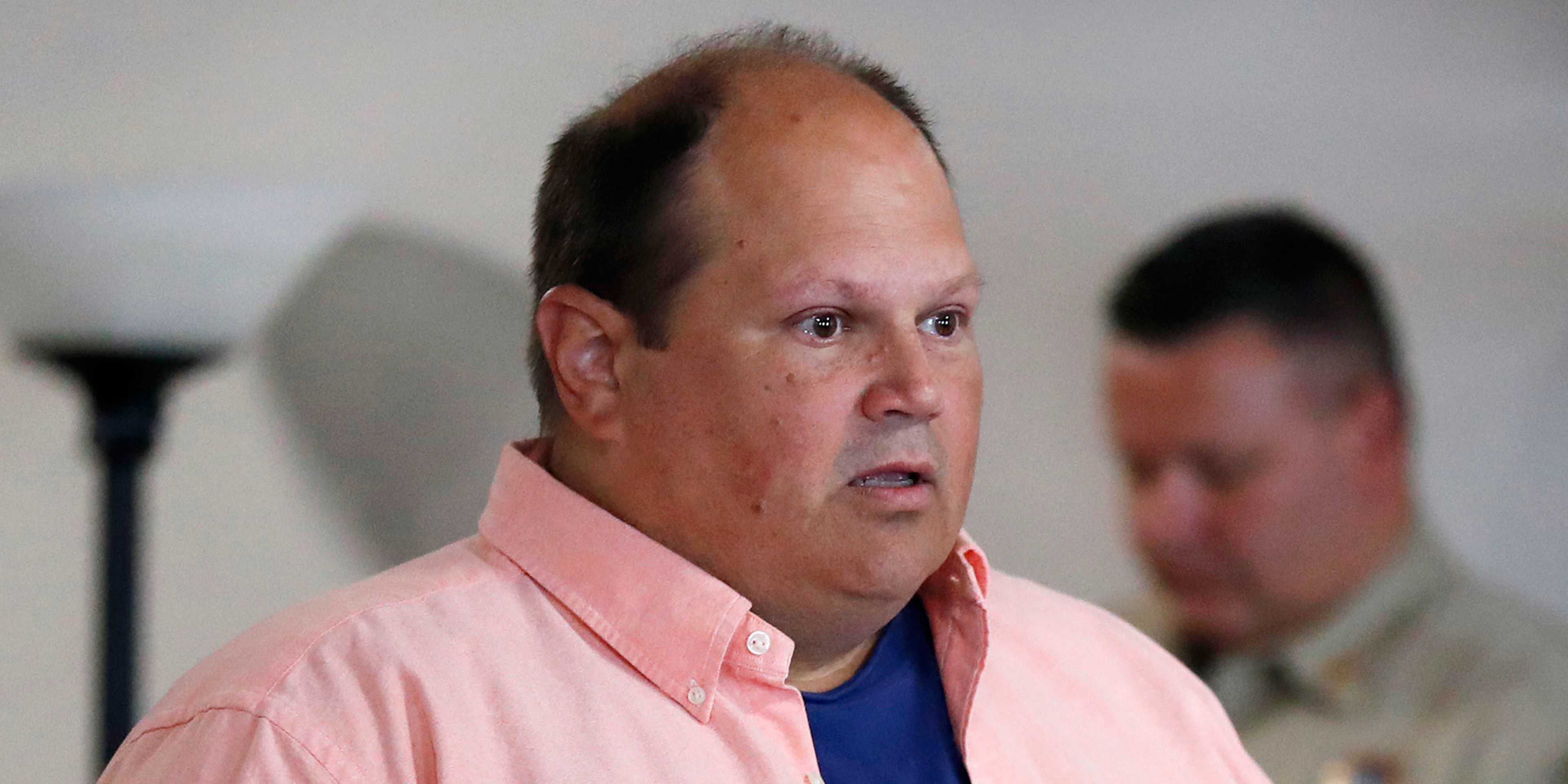 The image shows a middle-aged man wearing a pink shirt, with a serious expression on his face, standing in what appears to be an indoor setting.