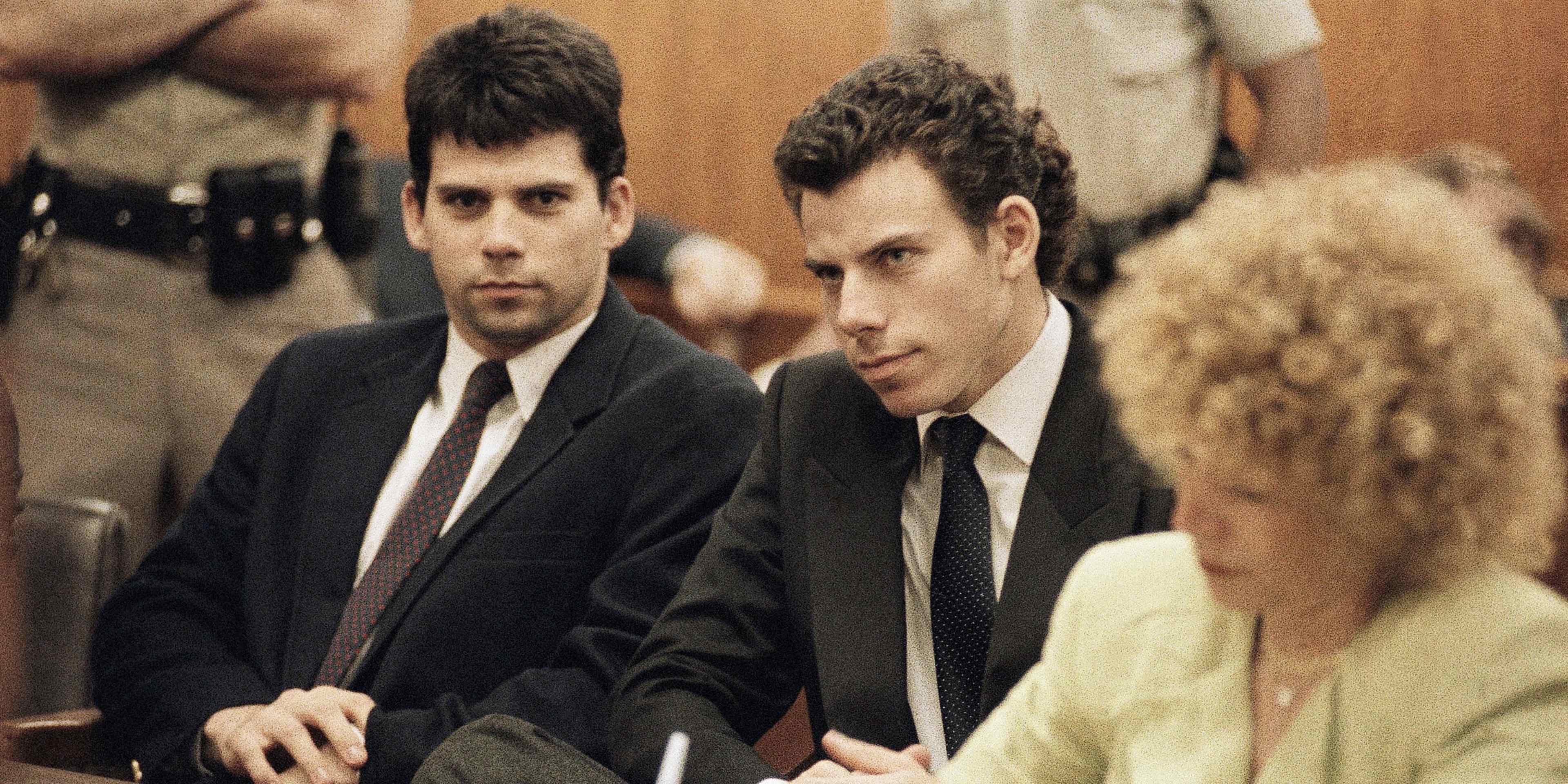 Two men in suits sitting at a table, with a woman in the background.