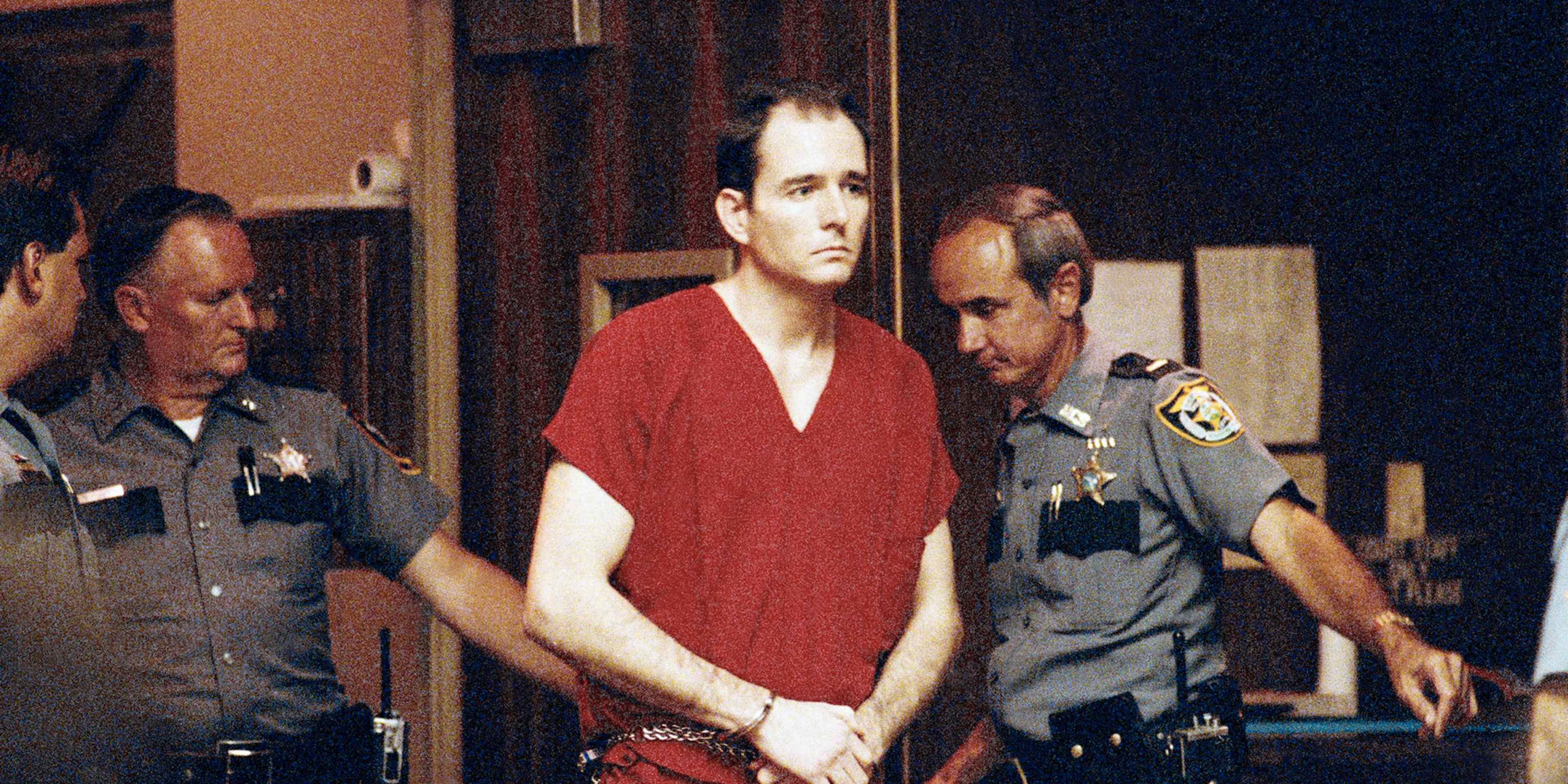 The image shows a man in a red robe being escorted by two police officers in a dimly lit setting.