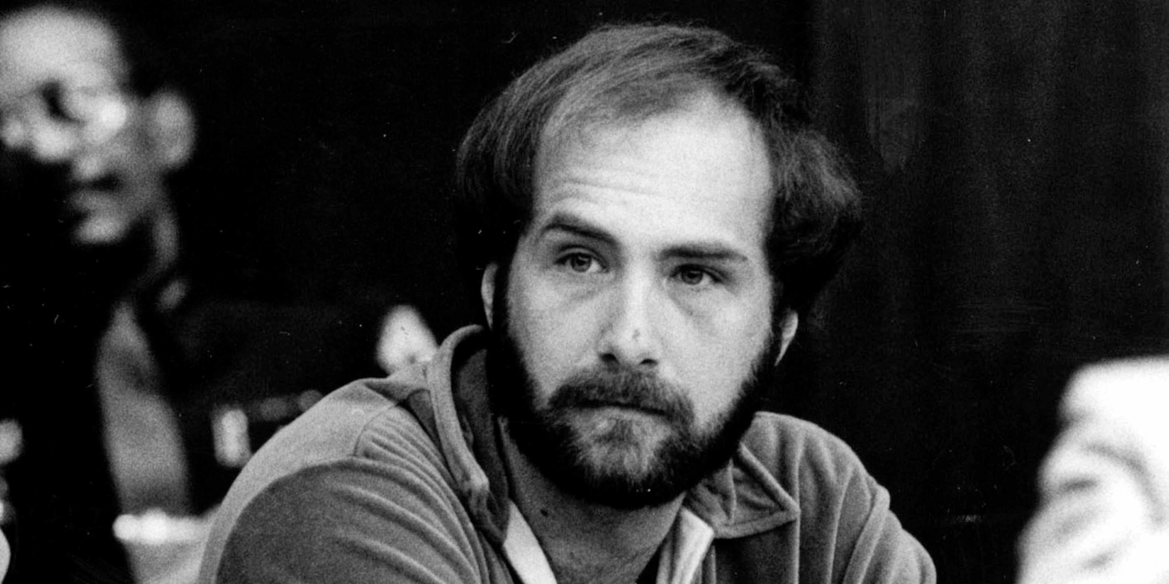 A middle-aged man with a beard and long hair, wearing a casual shirt, appears to be in a dimly lit setting with a dark background.