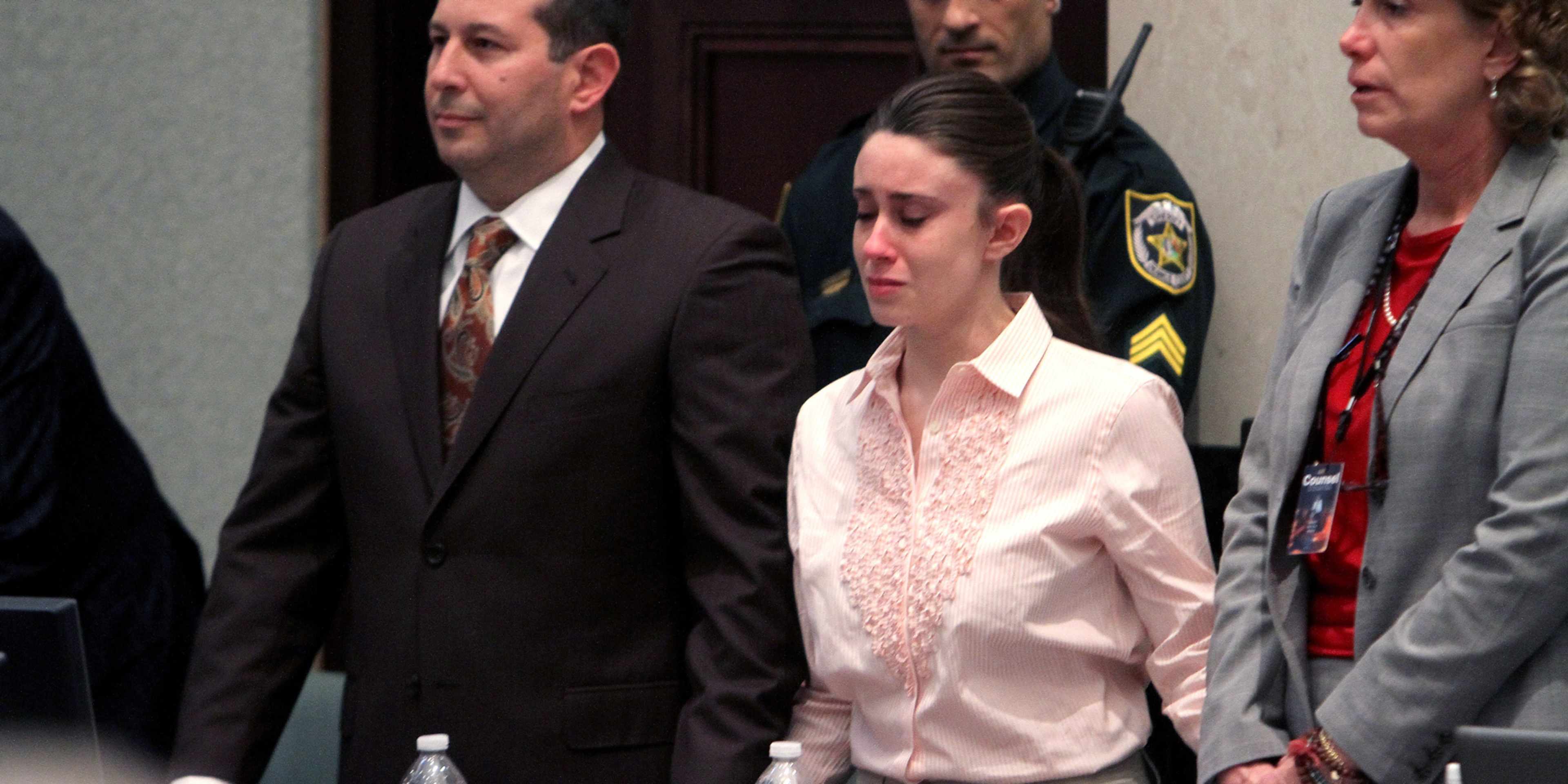 The image shows several individuals, including a woman in a pink shirt, walking together in what appears to be a formal setting, such as a courtroom or government building.