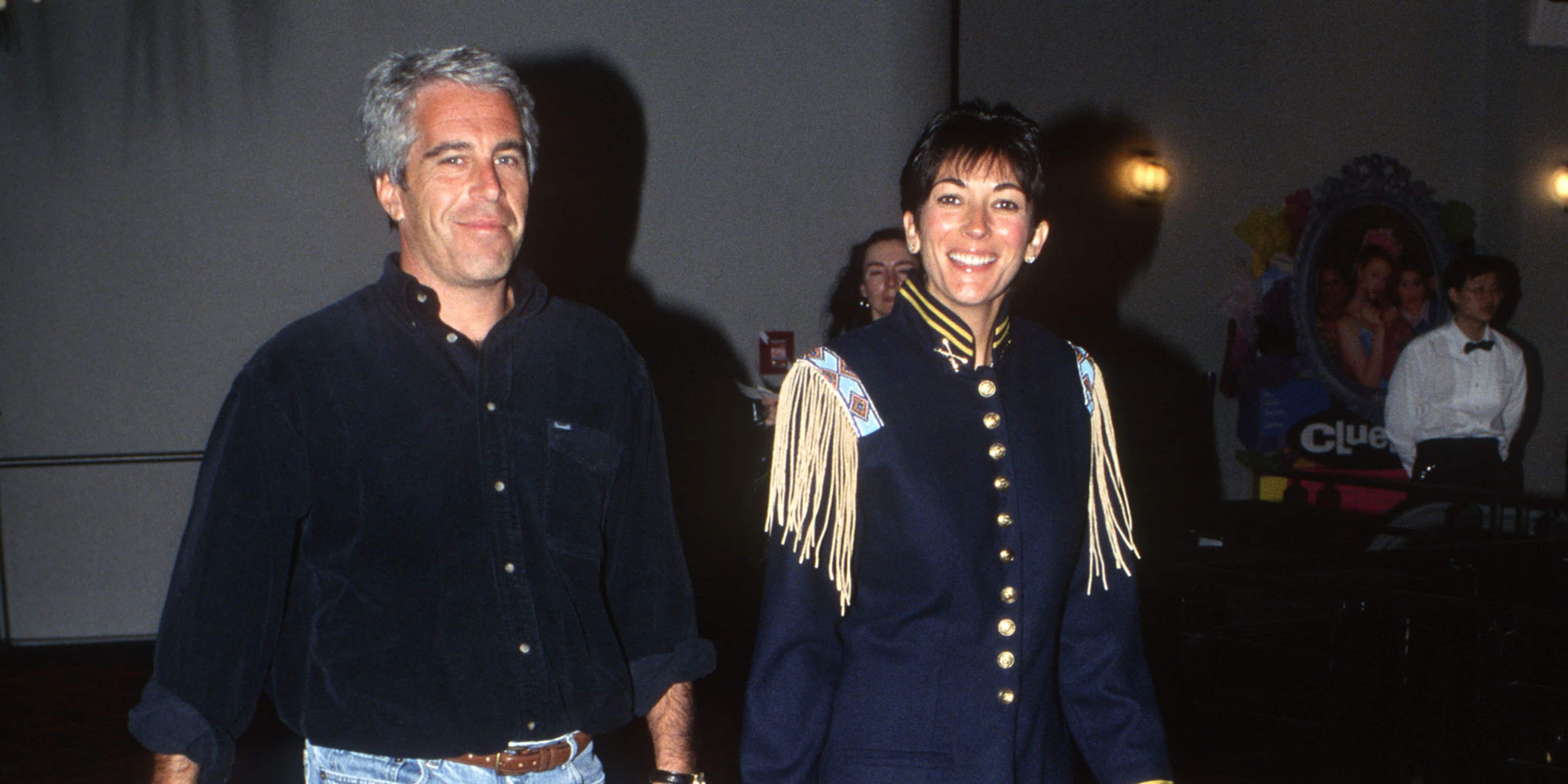 The image shows two individuals, a man and a woman, standing together in what appears to be a formal setting, with the woman wearing a military-style jacket and the man dressed casually in jeans and a jacket.